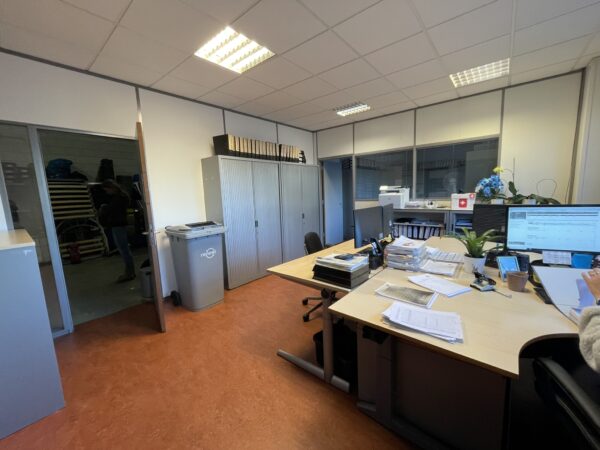 Kantoorruimte met bureaus, documenten, computerschermen en een open deur naar een magazijnruimte.