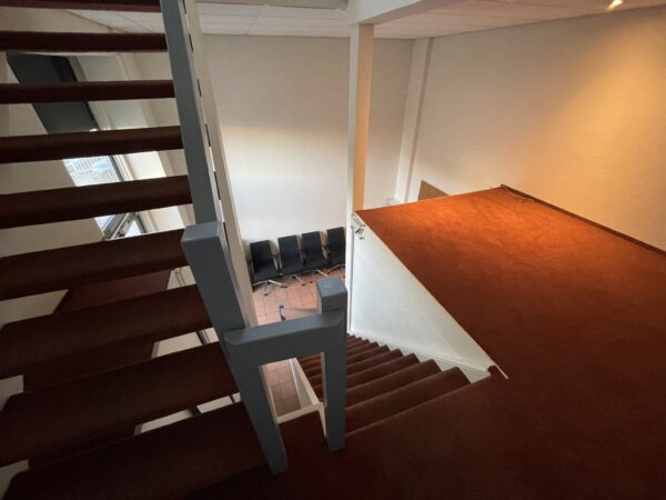 Trap met rood tapijt in een split-level ruimte met witte muren en zwarte wachtstoelen beneden.