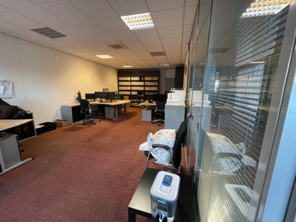 Kantoorruimte met meerdere bureaus, computers, archiefkasten en een glazen tussenwand.