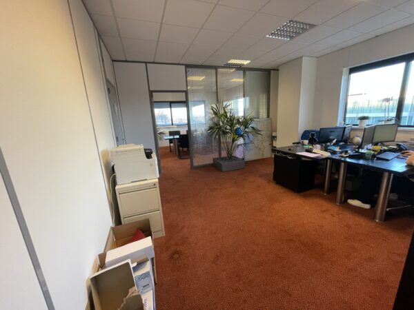 Kantoorruimte met roodtapijt, werkplekken, printer en zicht op vergaderruimte achter glazen wand.