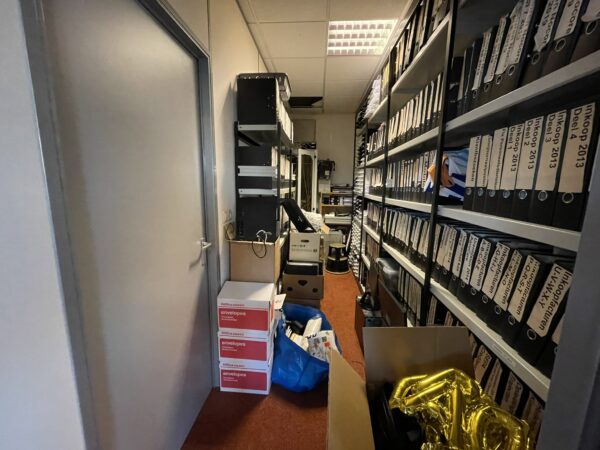 Volgepakte archiefruimte met rijen ordners, dozen en losse spullen op de vloer.