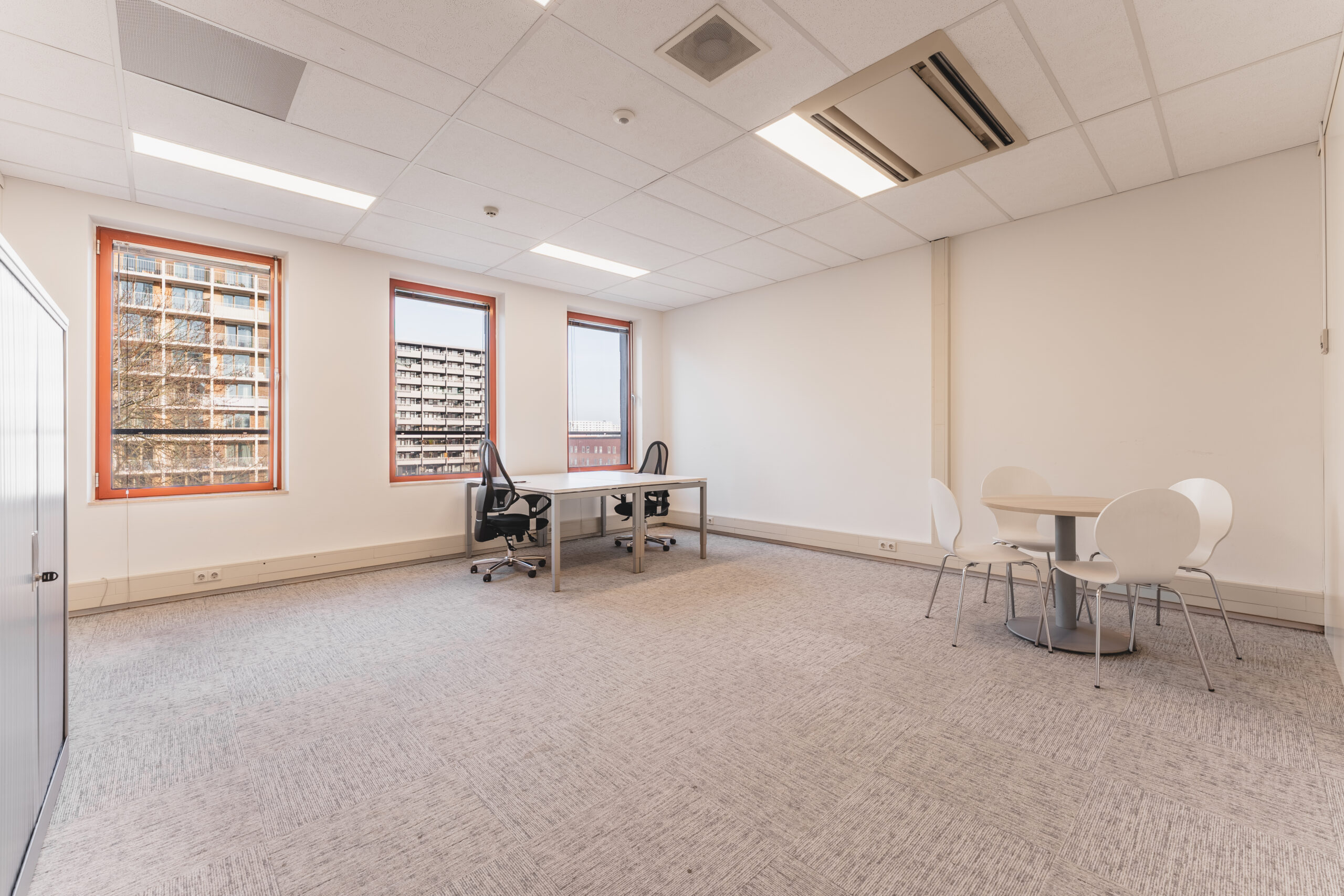 Een leeg kantoor met twee bureaus, bureaustoelen en een vergadertafel met stoelen.