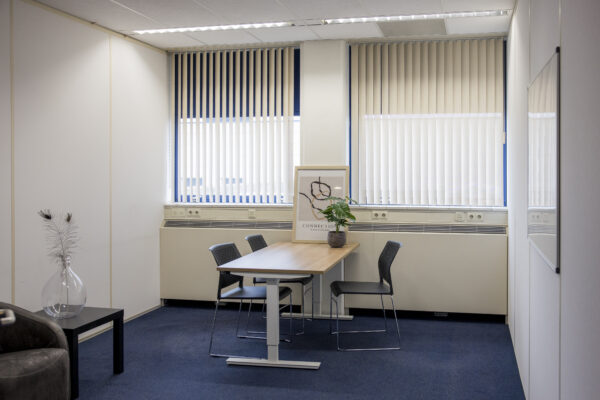 Een kantoor met een houten tafel, vier stoelen, een plant en een kunstwerk voor grote ramen met verticale jaloezieën.