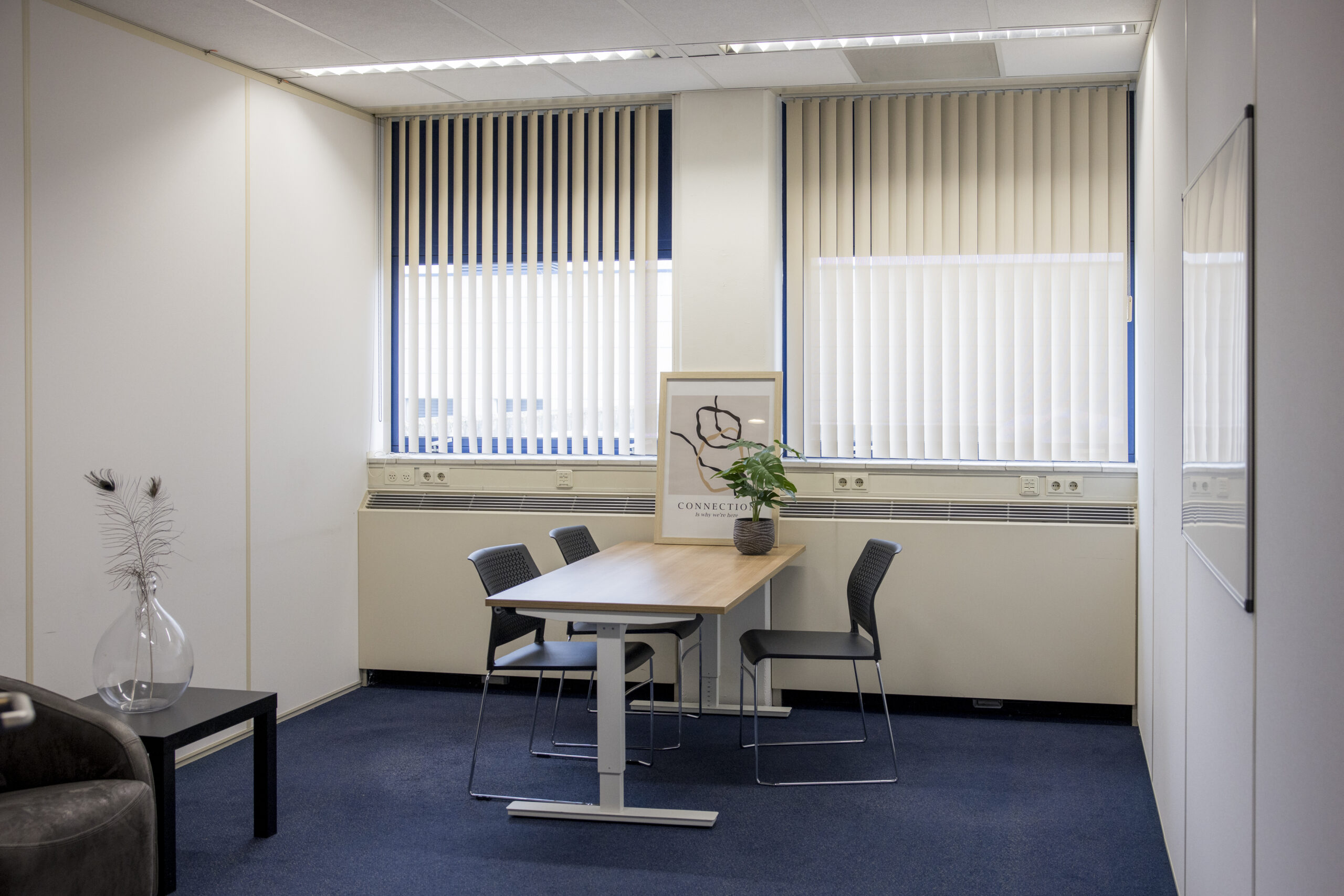 Een kantoor met een houten tafel, vier stoelen, een plant en een kunstwerk voor grote ramen met verticale jaloezieën.