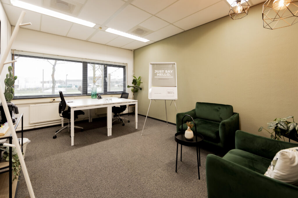 Een modern kantoor met groene fauteuils, een vergadertafel, en een flip-over met de tekst "Just Say Hello".