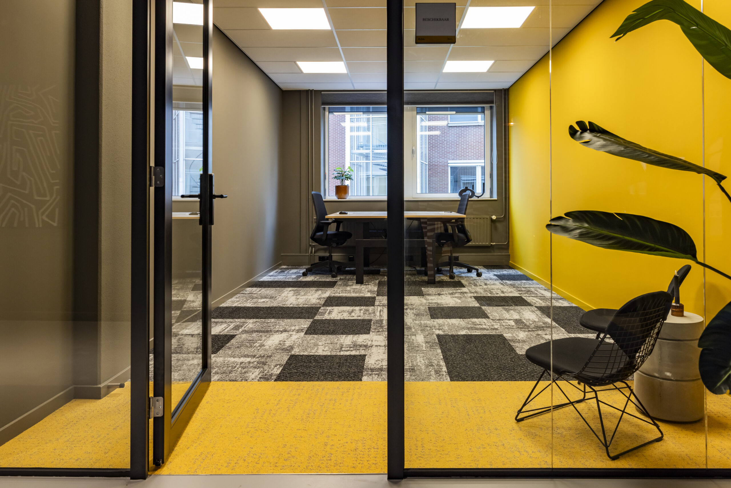 Een modern ingericht kantoor met een helder gele muur, glazen deur en een plant naast een bureau met twee stoelen.