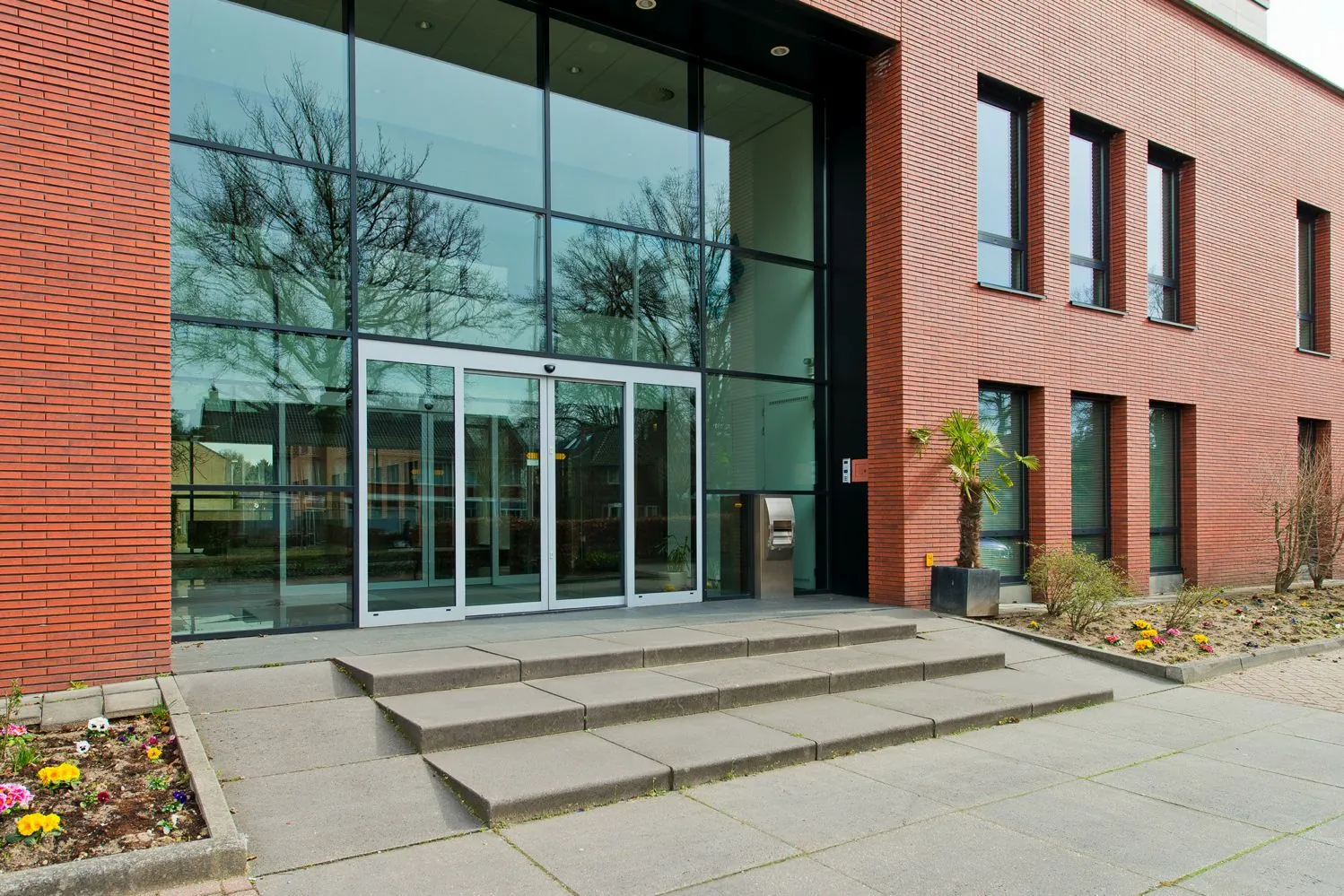 Ingang van een modern kantoorgebouw aan de Groenekanseweg met glazen schuifdeuren en een trap met bloemperken aan weerszijden.