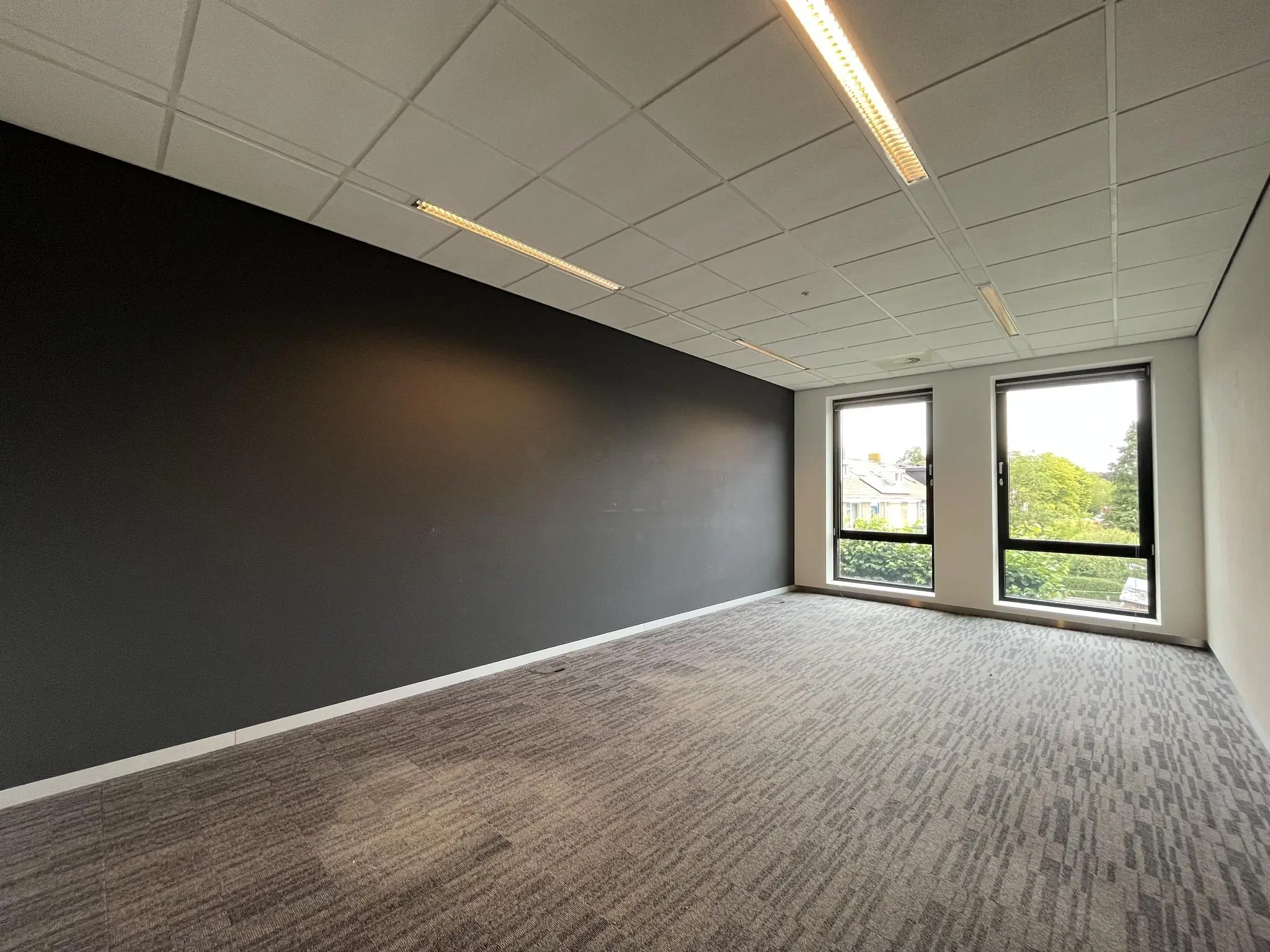 Leeg kantoor met grijze vloerbedekking, zwarte accentmuur en twee grote ramen aan de Groenekanseweg.