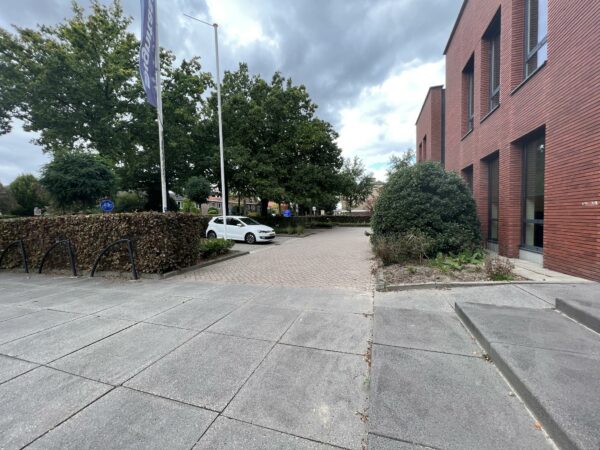 Ingang van een modern rood bakstenen gebouw aan de Groenekanseweg met parkeerplaatsen, een fietsenrek en bomen op de achtergrond.