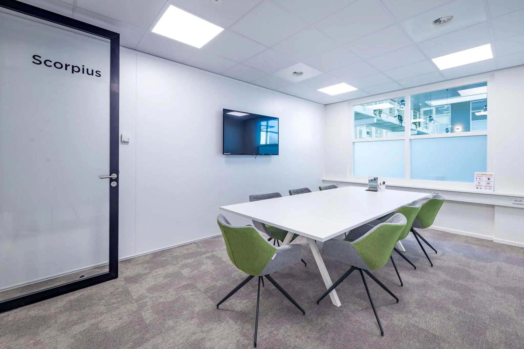 Vergaderruimte Scorpius met een witte tafel, zes groen-grijze stoelen, flatscreen aan de muur en groot raam met zicht op een modern kantoorgebouw.