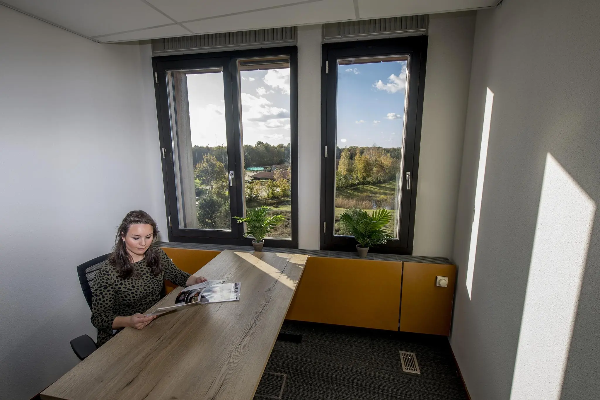 Vrouw leest een tijdschrift aan een bureau in een kantoorruimte aan de Fokkerstraat met uitzicht op een groene omgeving.