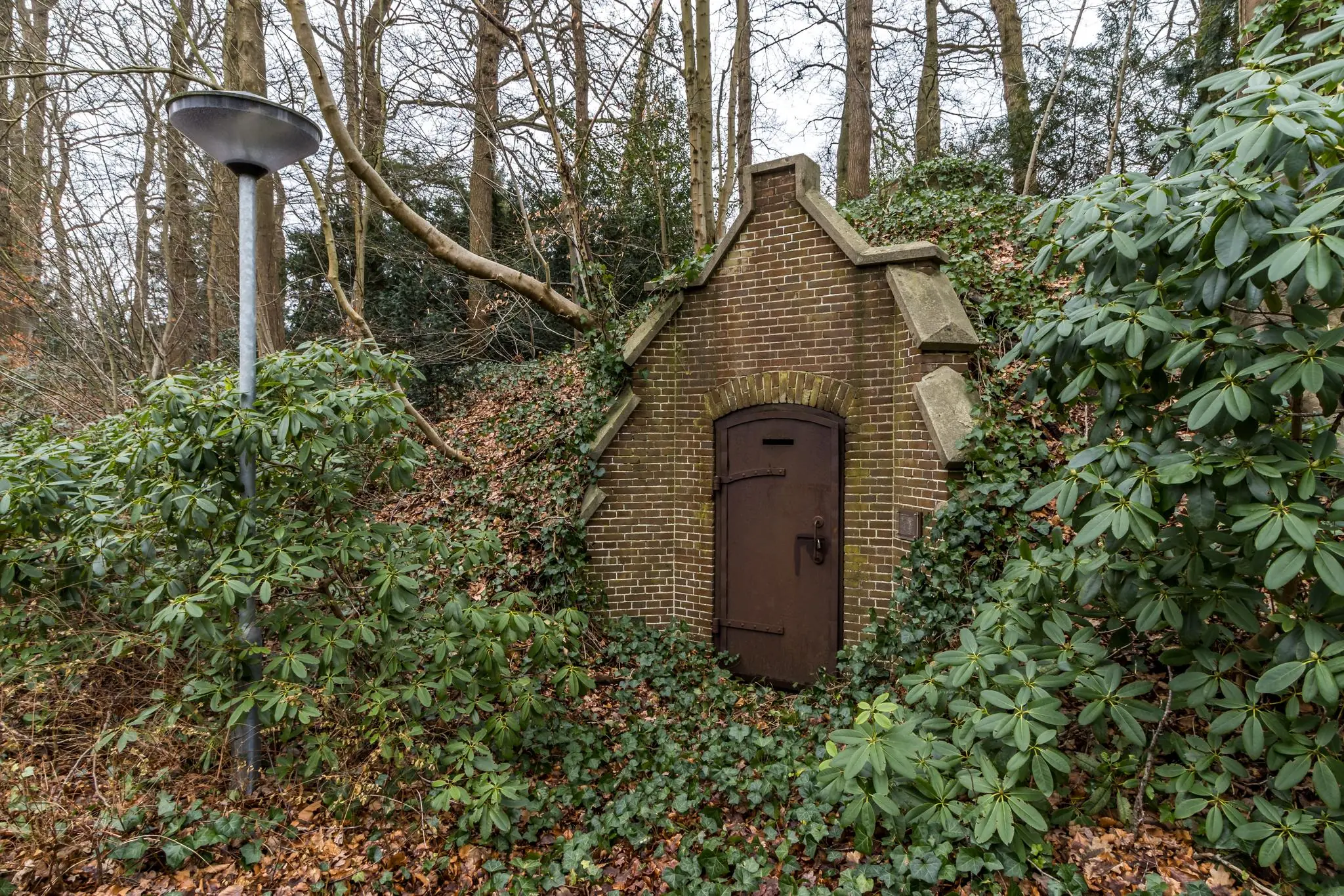 Bakstenen toegangspoort in een met klimop begroeide heuvel aan de Driebergseweg, omringd door bomen en struiken.