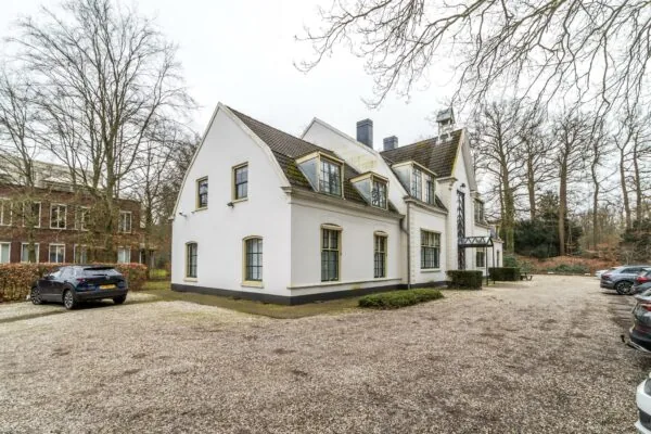 Wit landhuis aan de Driebergseweg met parkeerplaatsen op een grindterrein en omliggende bomen.