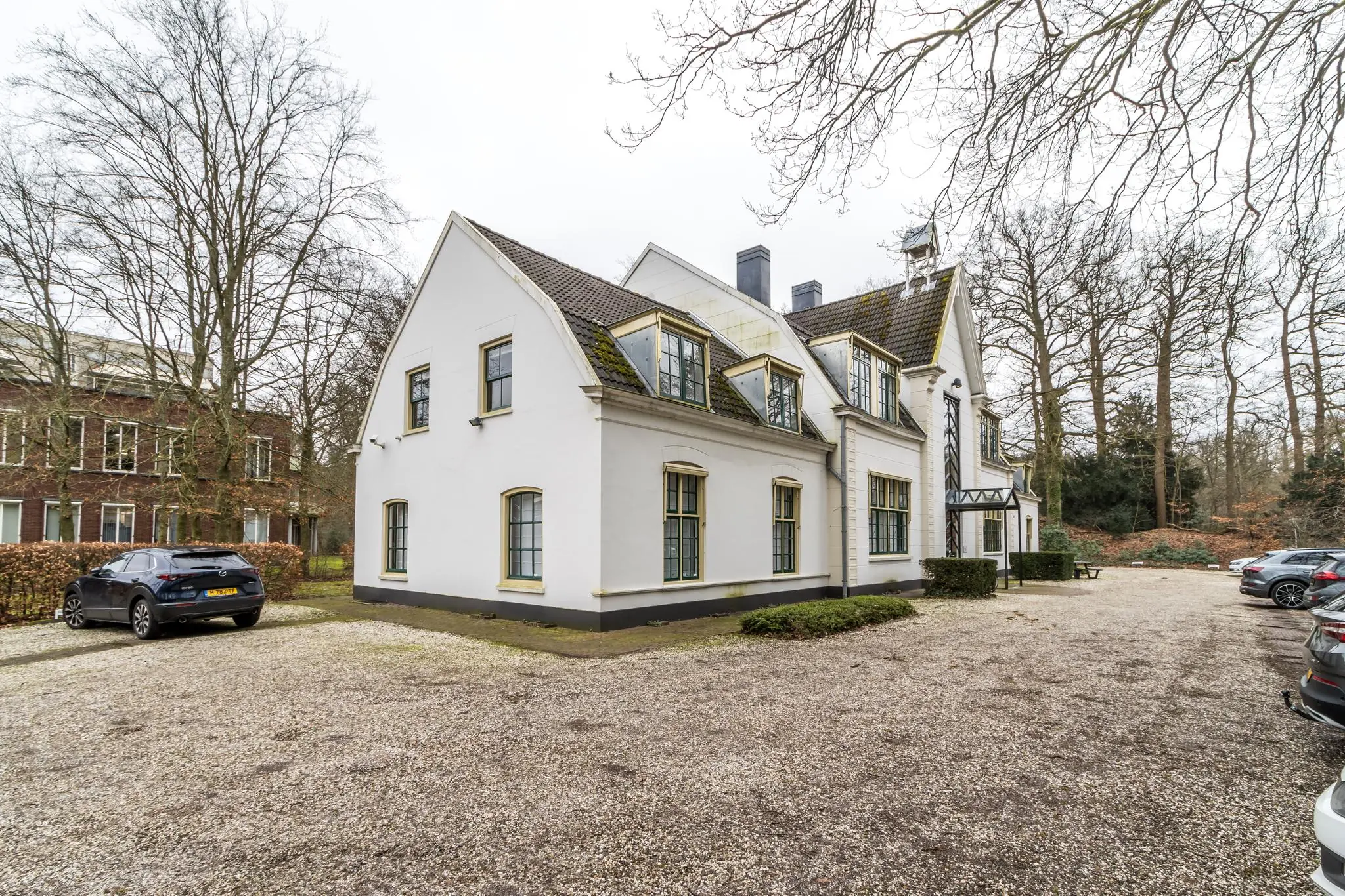 Wit landhuis aan de Driebergseweg met parkeerplaatsen op een grindterrein en omliggende bomen.