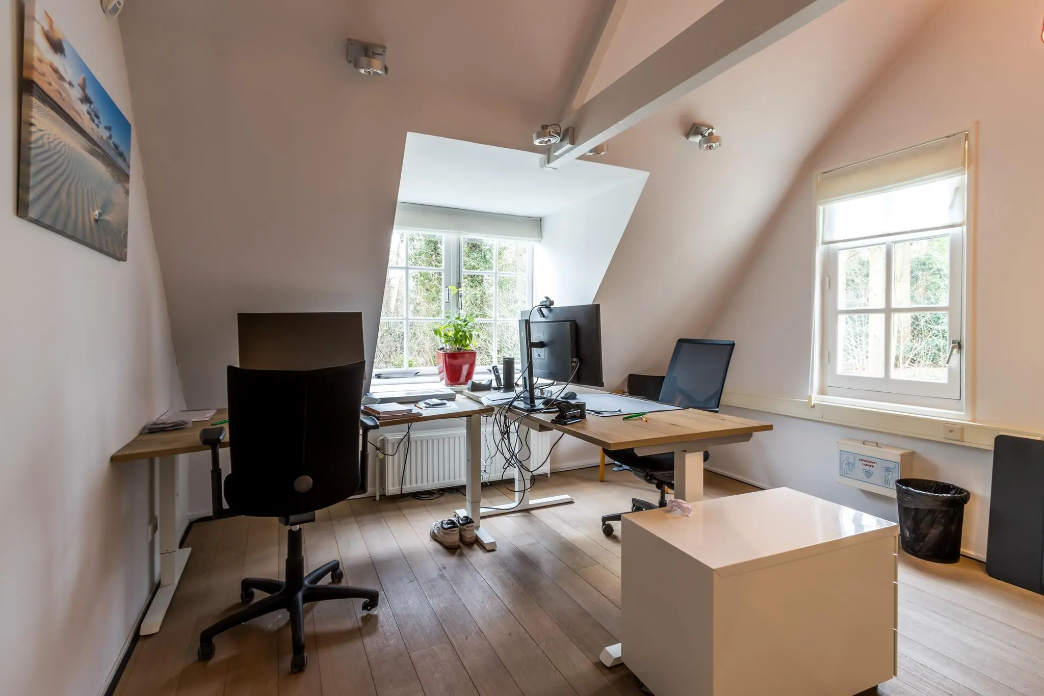 Zolderkantoor aan de Driebergseweg met twee bureaus, computers en een raam met uitzicht op bomen.