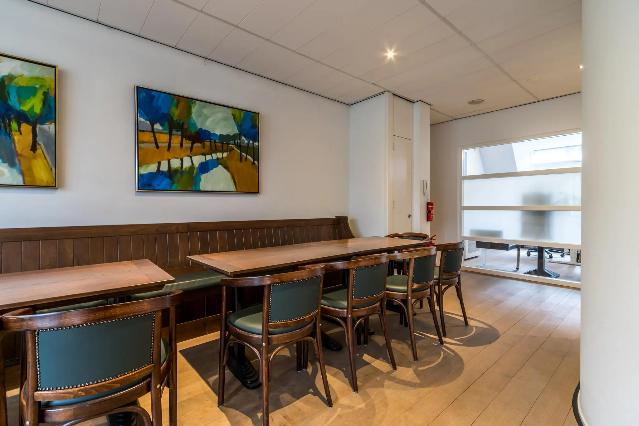 Vergaderruimte aan de Driebergseweg met houten tafel, groene stoelen en kleurrijke schilderijen aan de muur.