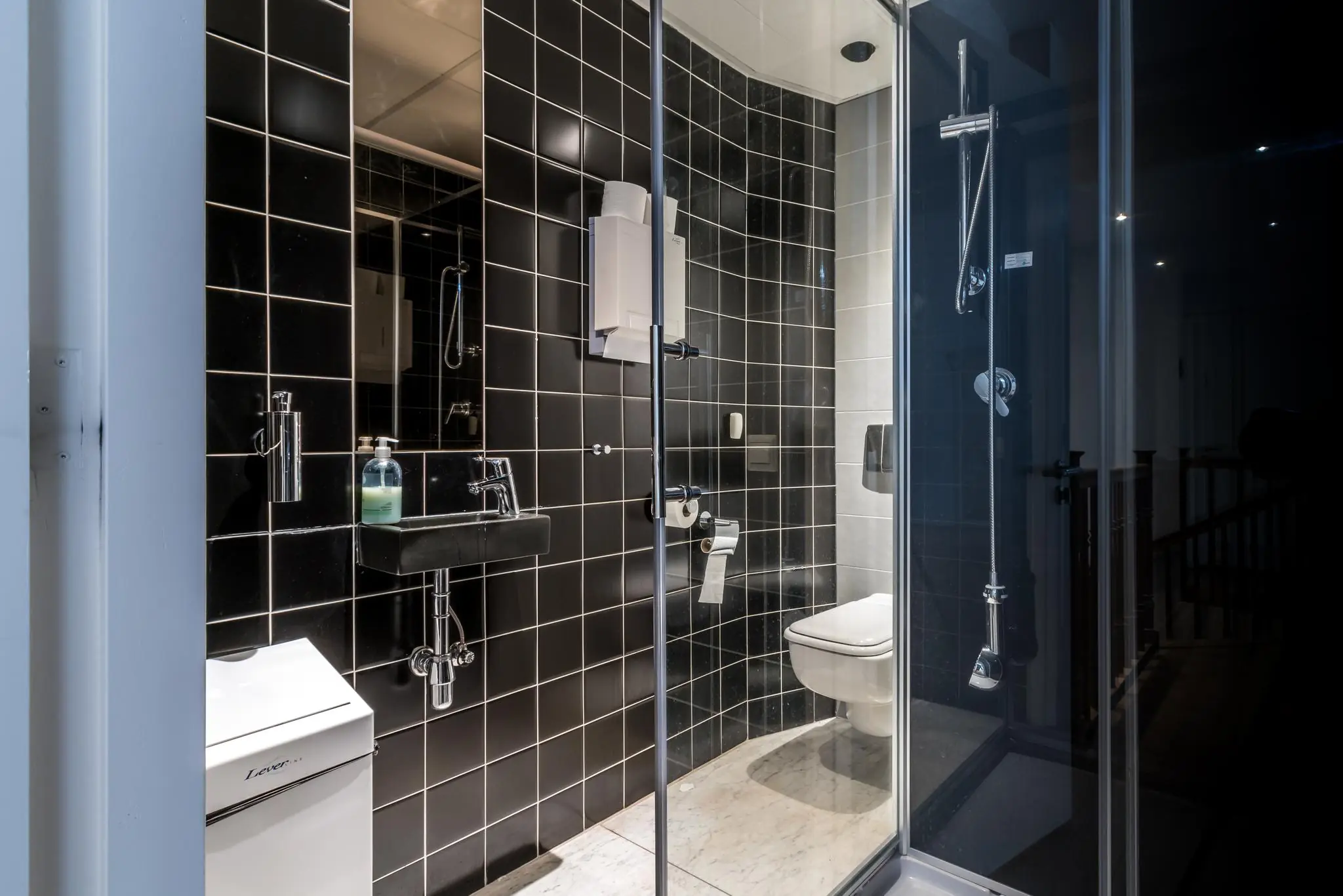 Moderne badkamer met zwarte wandtegels, een glazen douchecabine, zwevend toilet en een zwarte wastafel.