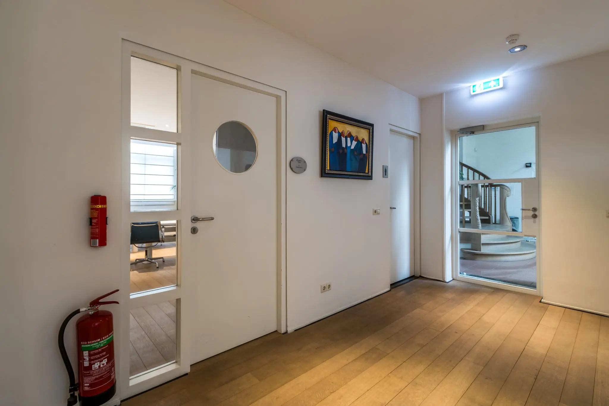 Moderne hal met houten vloer, brandblusser, kunst aan de muur en zicht op een trap via een glazen deur in een kantoorpand aan de Driebergseweg.