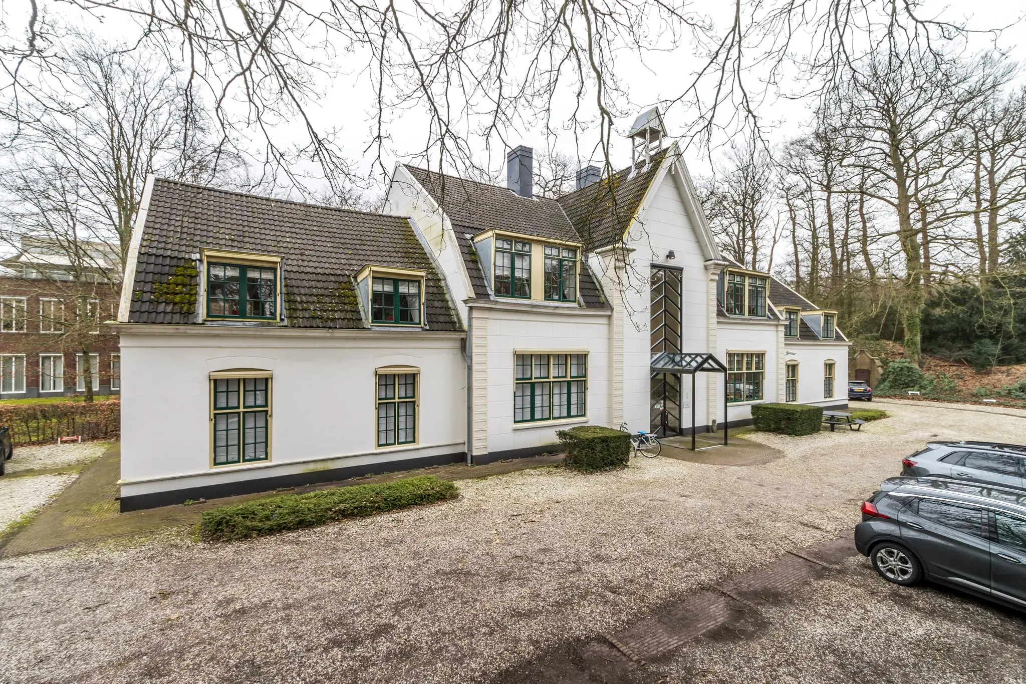 Wit herenhuis aan de Driebergseweg met donkere dakpannen, omringd door geparkeerde auto's en bomen.