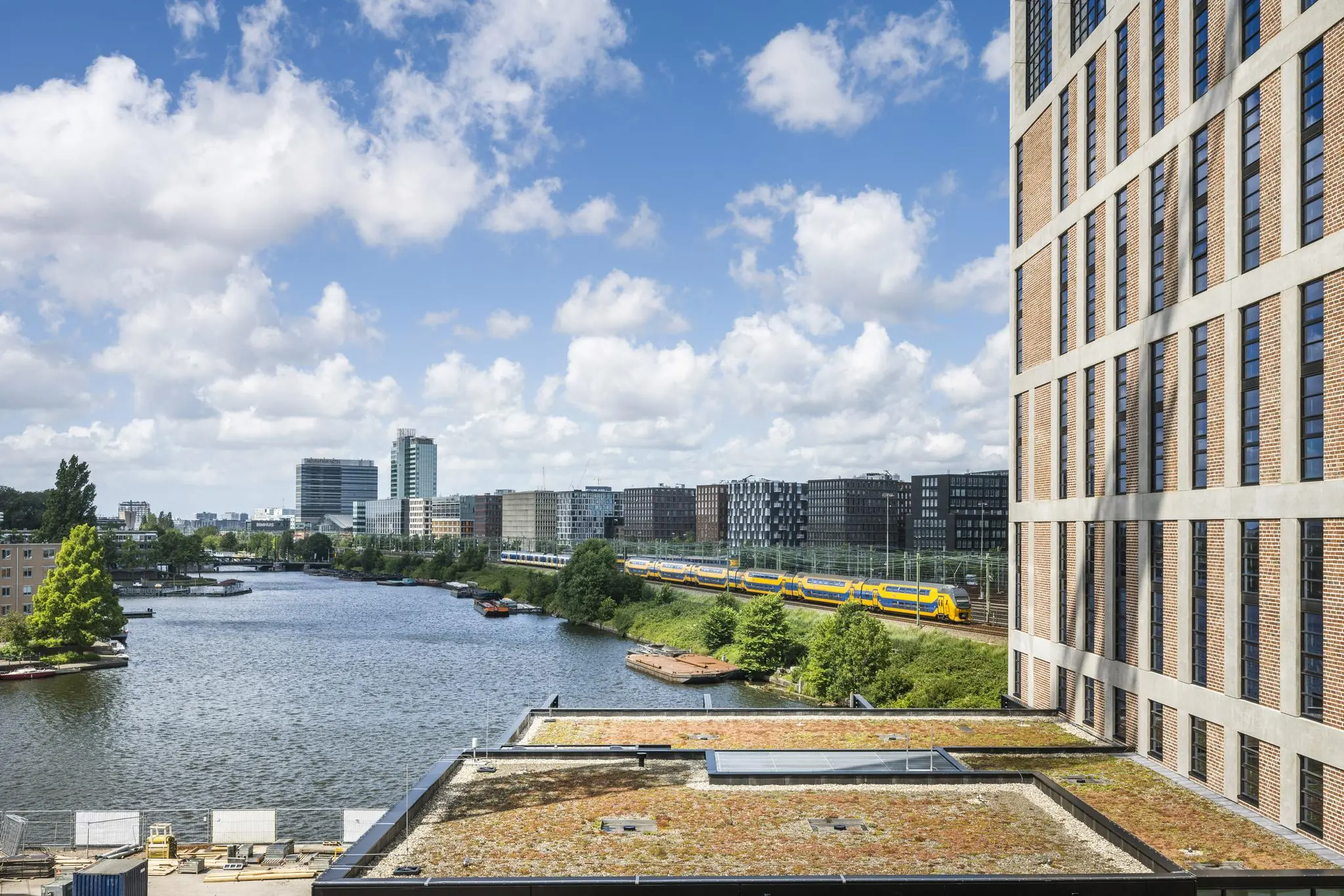 Uitzicht op de Pieter Goosstraat in Amsterdam met een geel-blauwe trein langs het water, omringd door moderne gebouwen en een wolkenlucht.