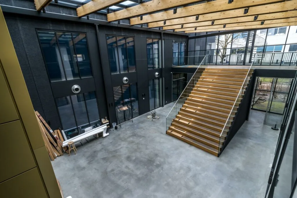 Ruime, moderne hal aan de Laarderhoogtweg met een brede houten trap, glazen balustrades en zichtbare houten balken in het plafond.