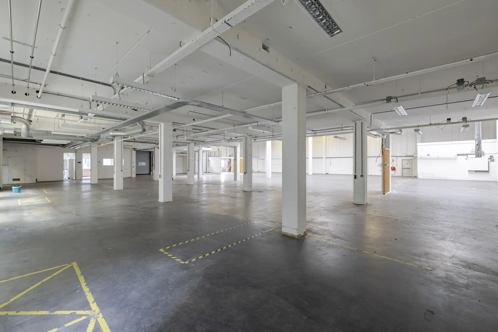 Ruime, lege industriële loods aan de Laarderhoogtweg met betonnen vloer, witte steunpilaren en zichtbare leidingconstructies aan het plafond.