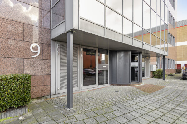 Ingang van een modern kantoorpand aan de Curieweg 9 met een draaiende toegangsdeur en spiegelende gevel.