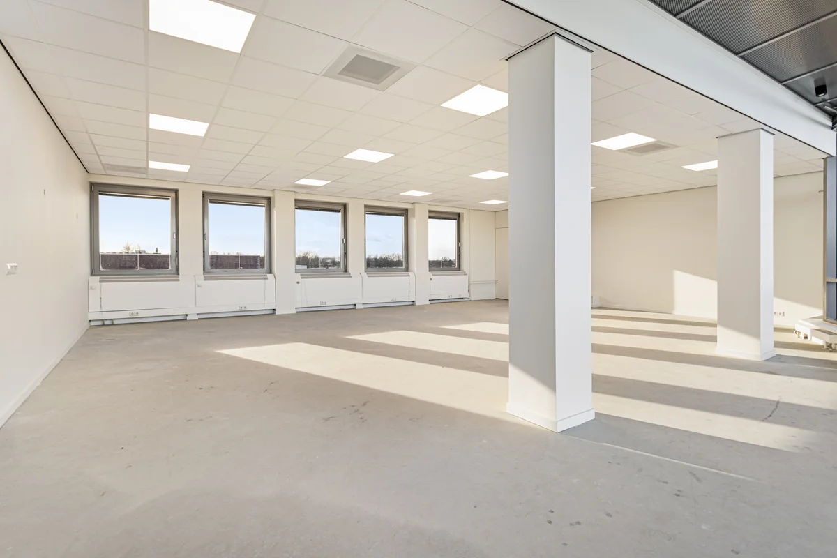 Lichte, lege kantoorruimte aan de Curieweg met grote ramen en witte plafonds met inbouwverlichting.