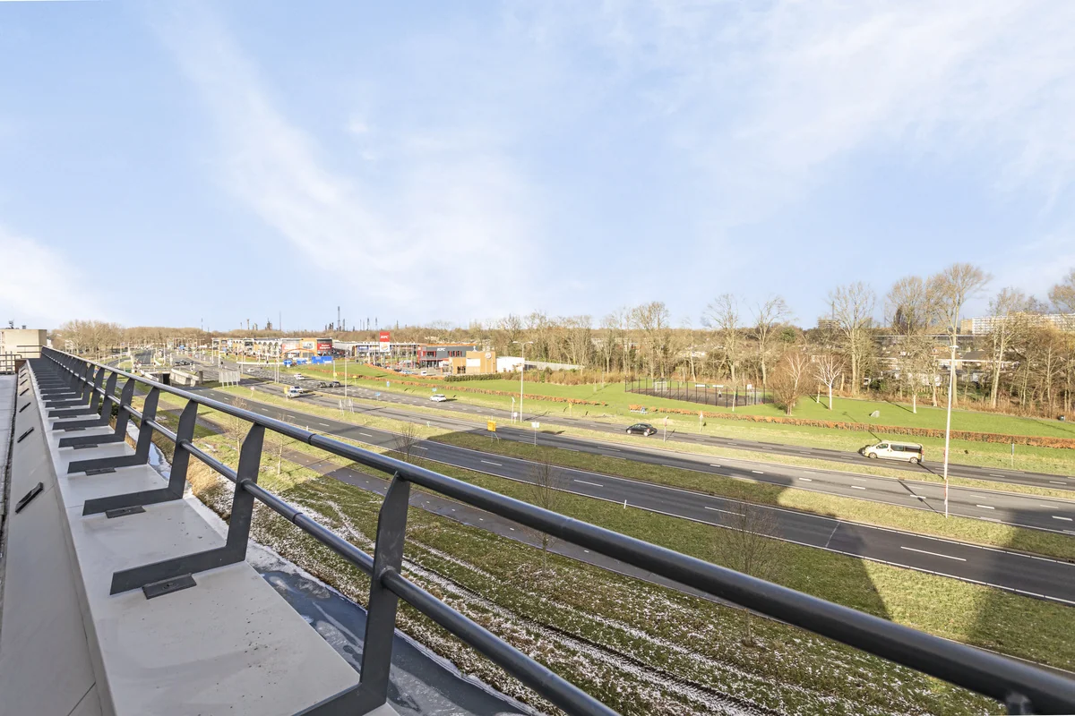 Uitzicht vanaf een balkon aan de Curieweg op een weg, groenstrook en nabijgelegen bedrijventerrein.