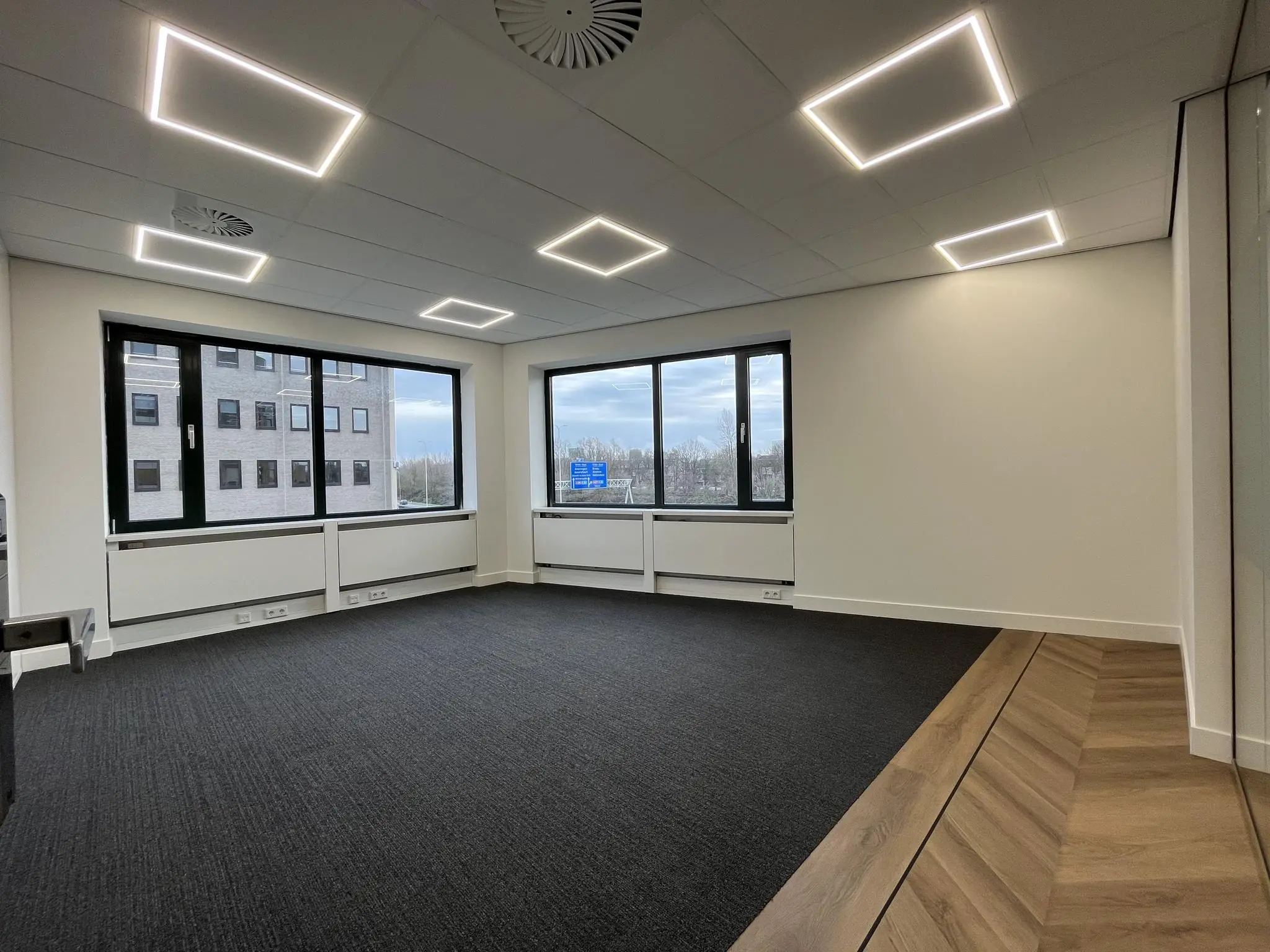 Modern, leeg kantoor aan de Euclideslaan met grote ramen, donker tapijt en ingebouwde LED-verlichting in het plafond.