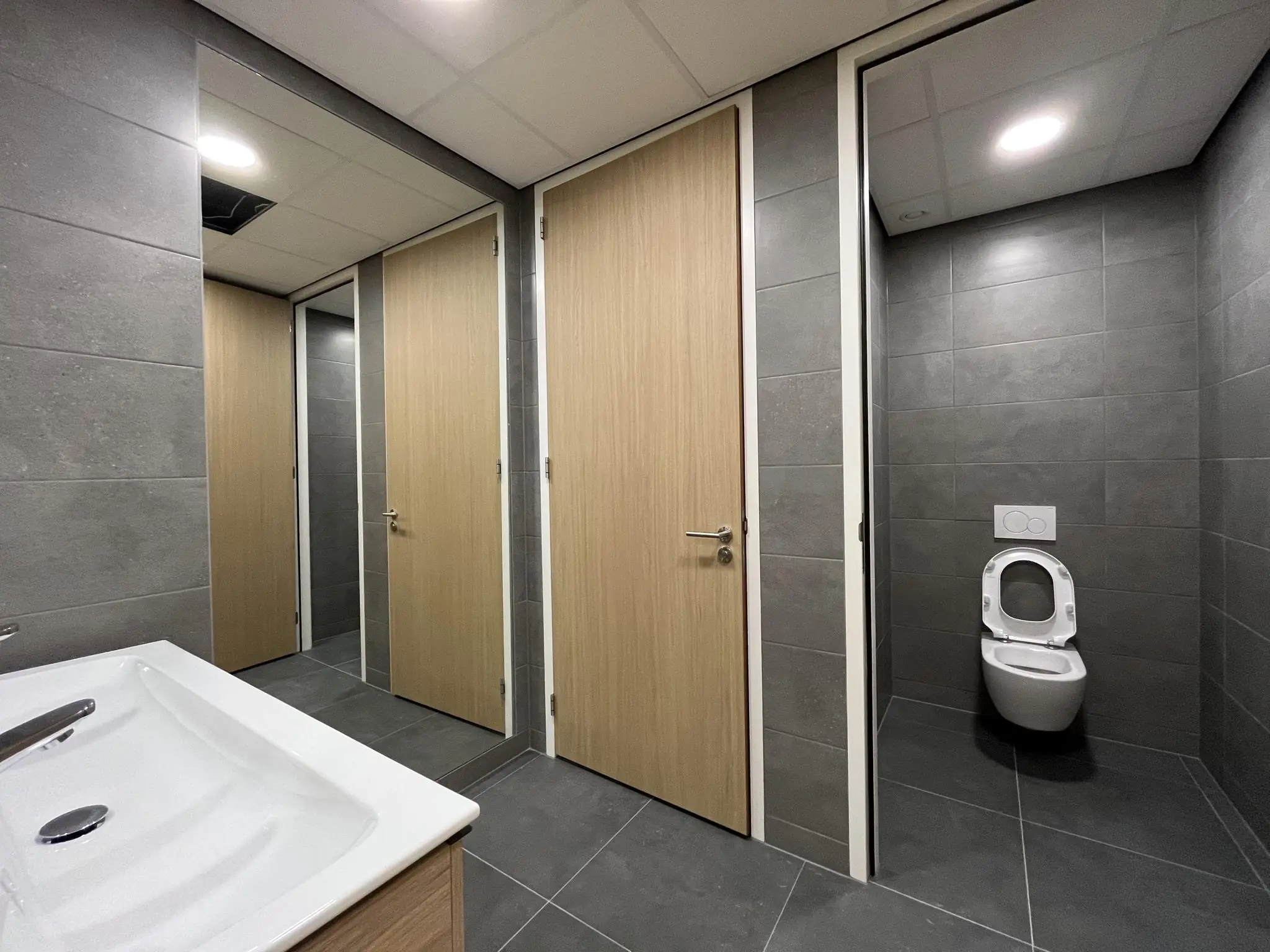 Moderne toiletruimte met grijze betegeling, houten deuren en een open toiletdeur waar een wandcloset zichtbaar is.