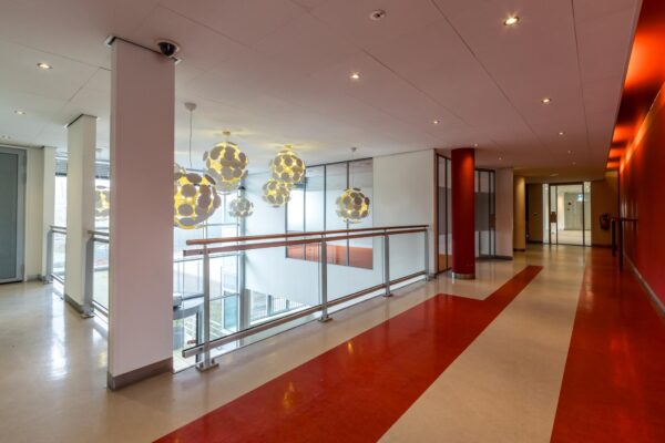 Moderne gang met rood-witte vloer, glazen balustrade en opvallende bolvormige hanglampen in een gebouw aan de Lemelerbergweg.