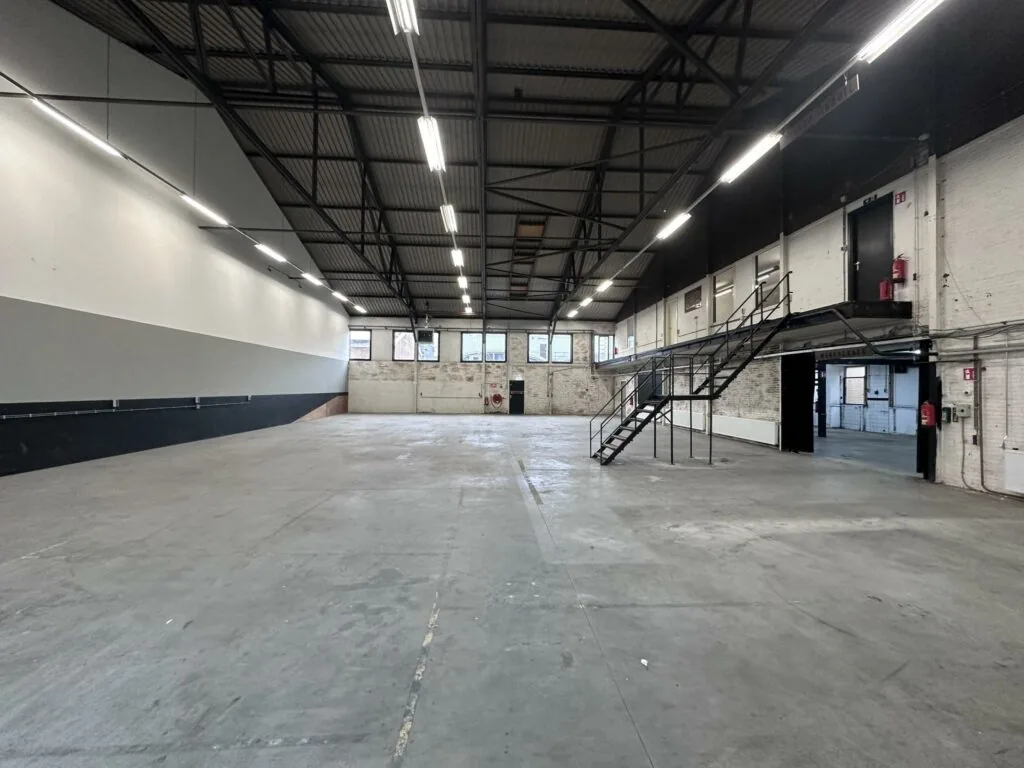 Ruime, lege industriële loods aan de Pilotenstraat met betonnen vloer, hoge plafonds en metalen trap naar een tussenverdieping.
