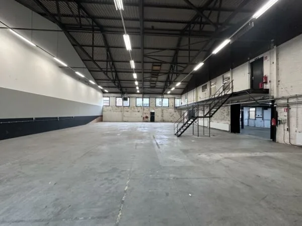 Ruime, lege industriële loods aan de Pilotenstraat met betonnen vloer, hoge plafonds en metalen trap naar een tussenverdieping.