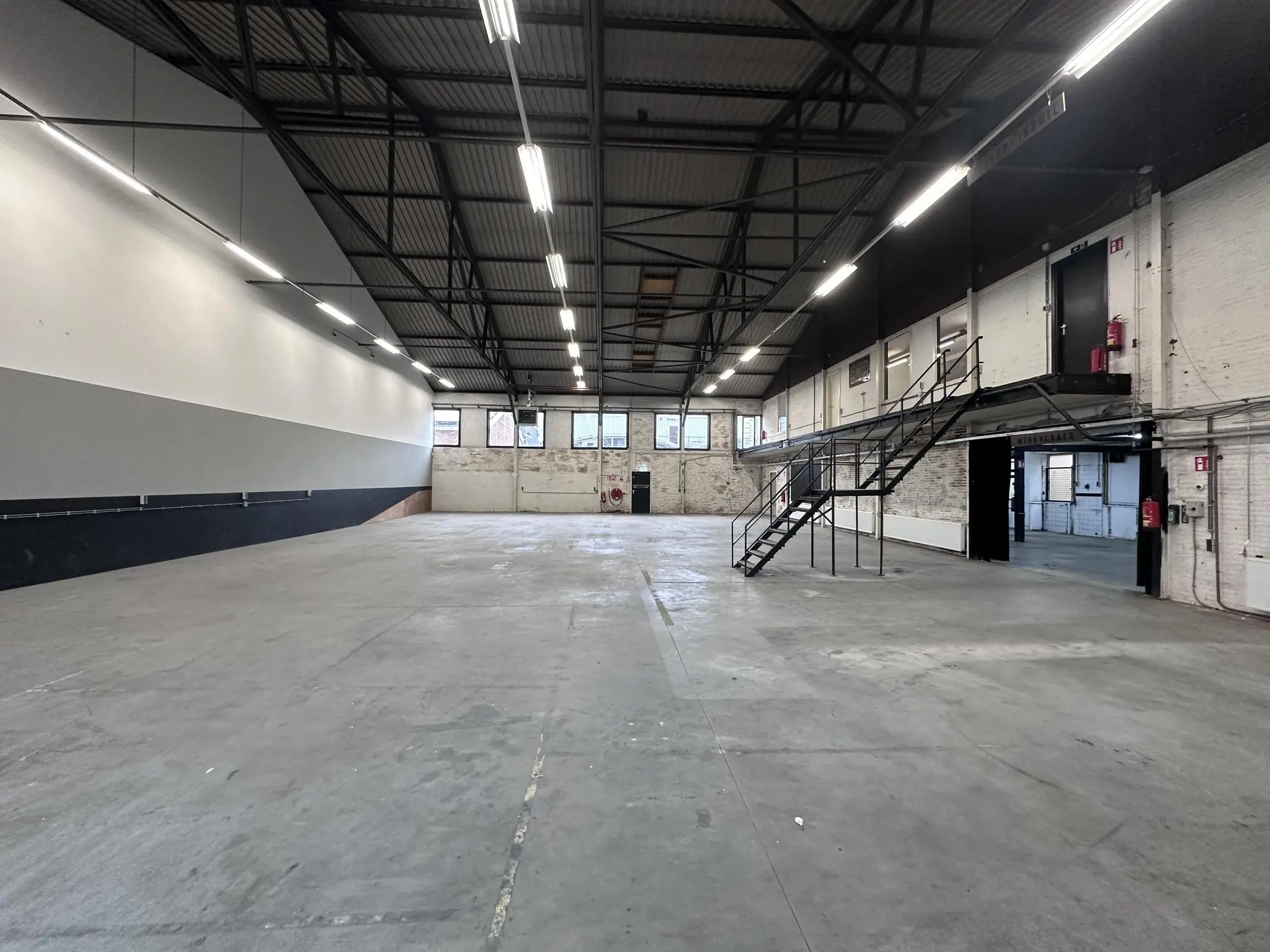 Ruime, lege industriële loods aan de Pilotenstraat met betonnen vloer, hoge plafonds en metalen trap naar een tussenverdieping.