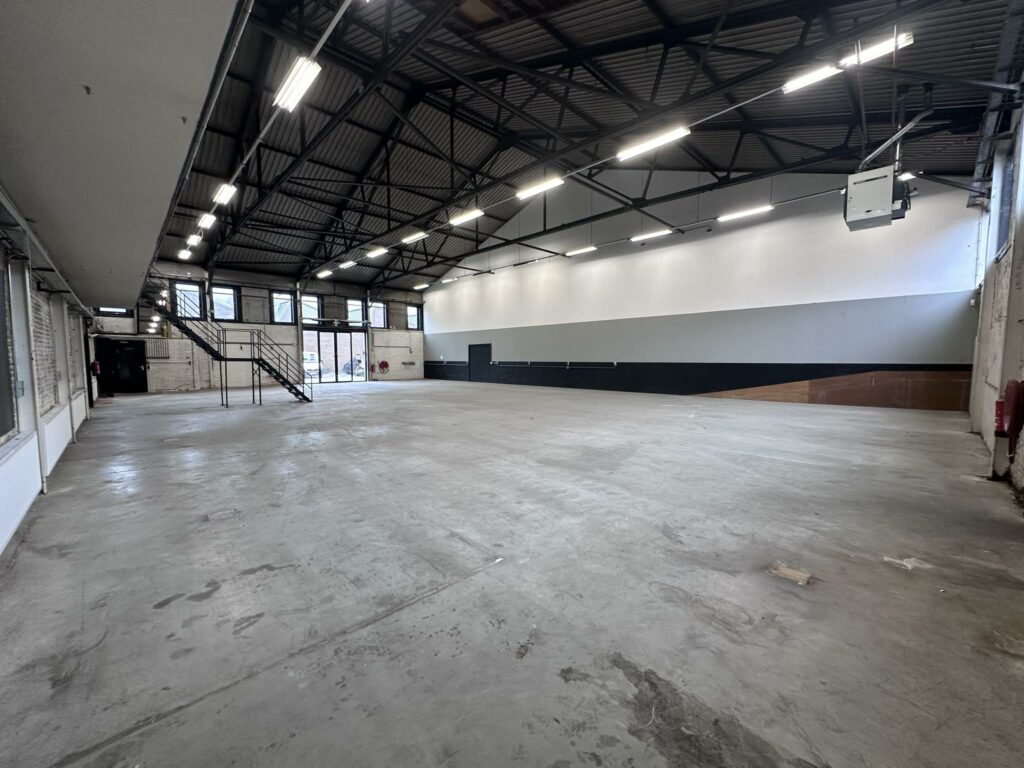 Ruime, lege bedrijfshal aan de Pilotenstraat met betonnen vloer, hoge plafonds en metalen trap.