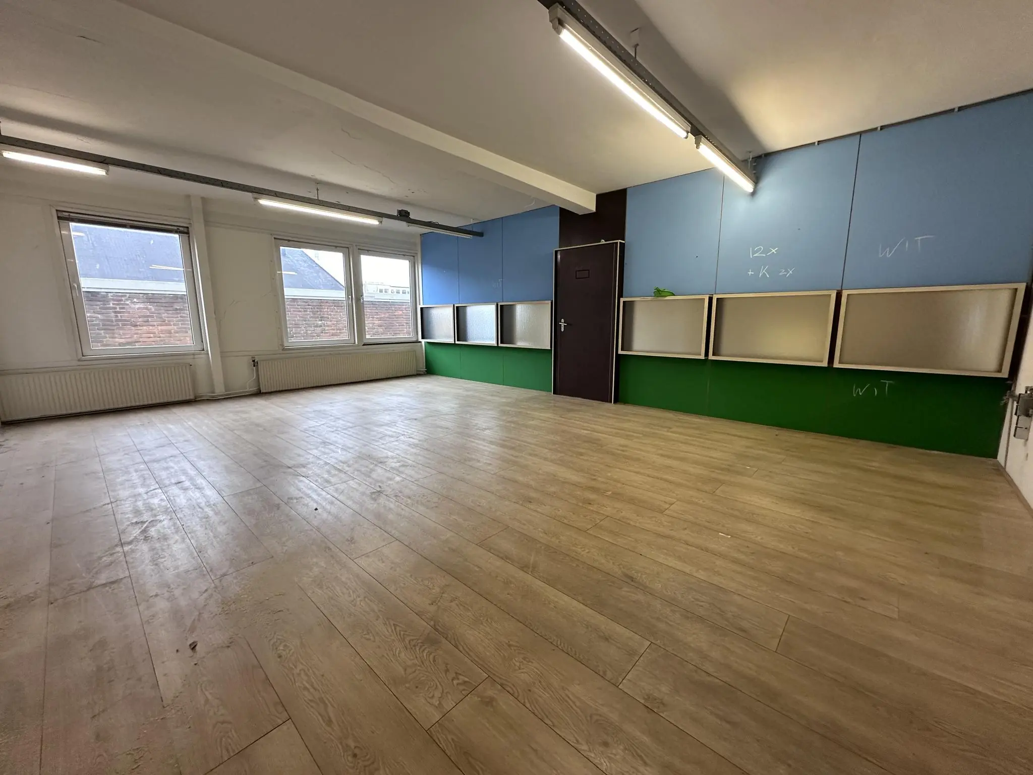 Leeg lokaal aan de Pilotenstraat met houten vloer, drie ramen en gekleurde muurpanelen met krijtaantekeningen.