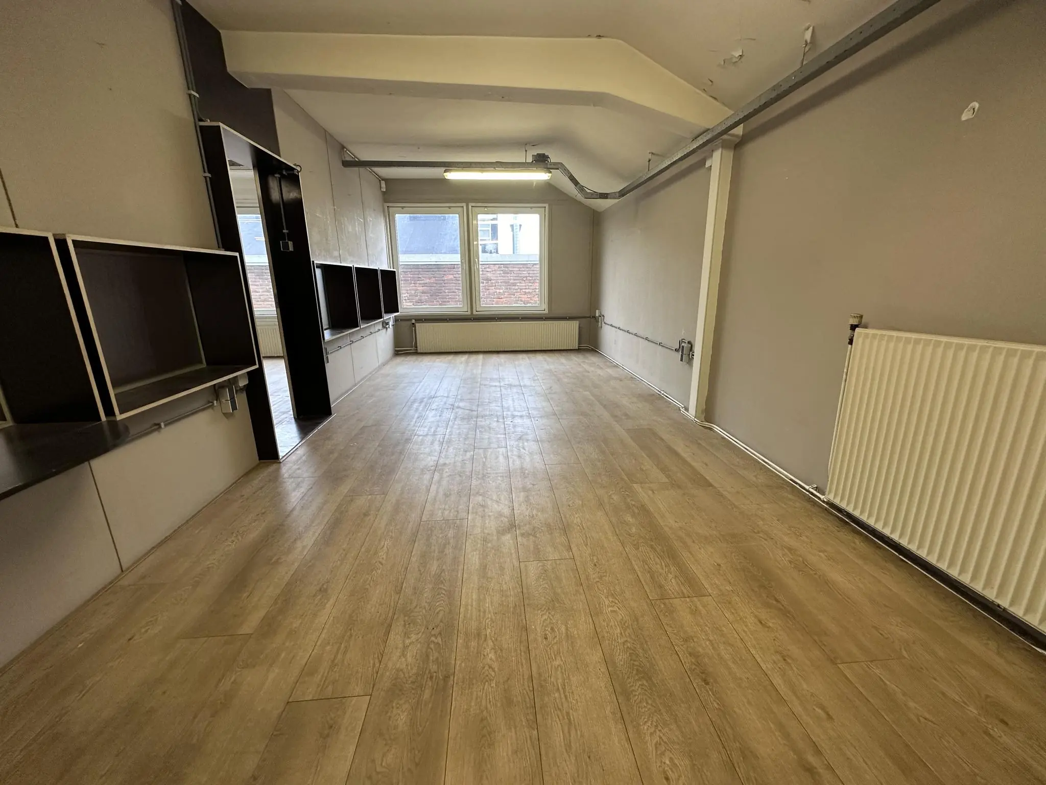 Lege kantoorruimte aan de Pilotenstraat met houten vloer, wandkasten en grote ramen aan het eind.