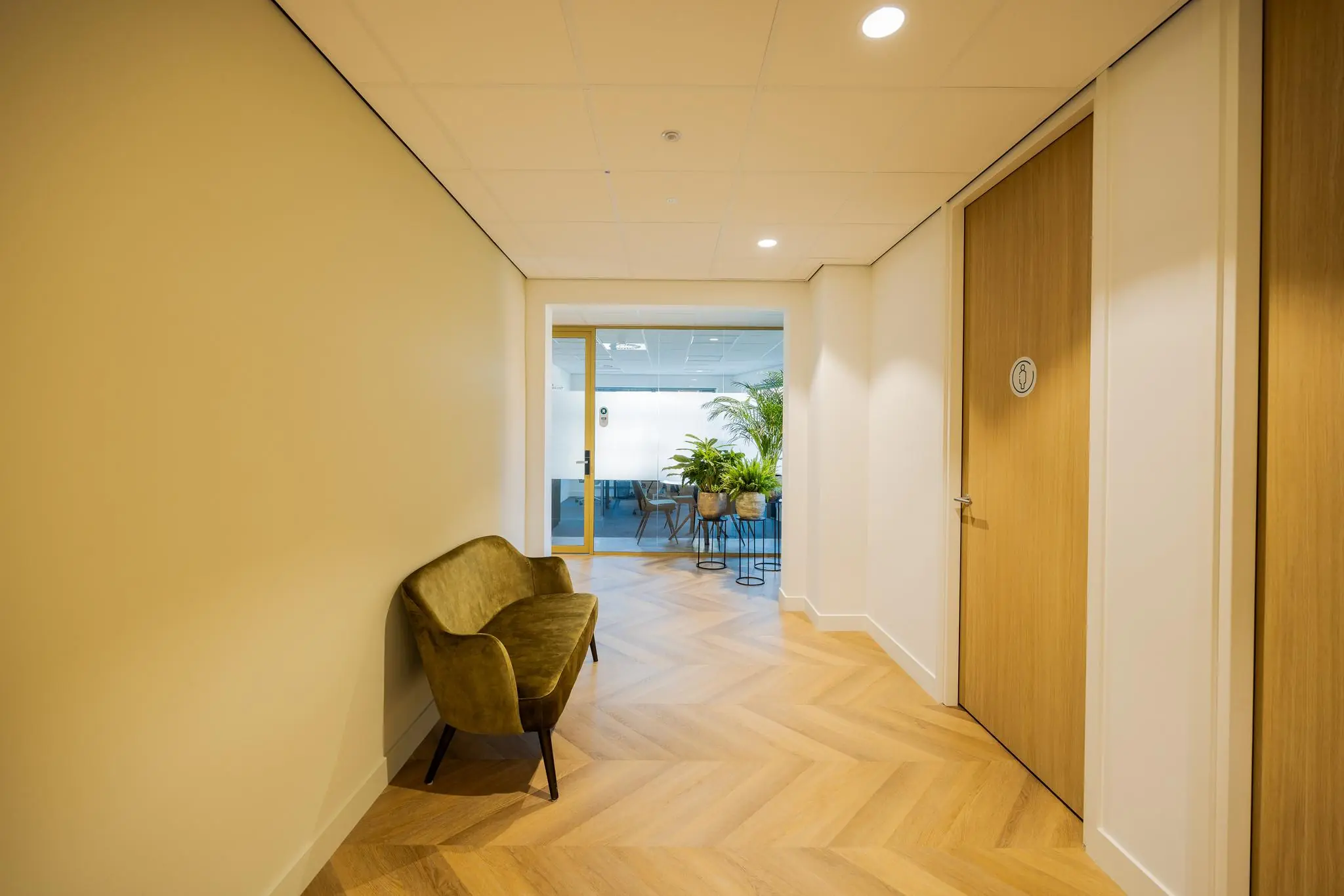 Hal met visgraatvloer, groen fluwelen bankje en uitzicht op vergaderruimte aan de Euclideslaan.