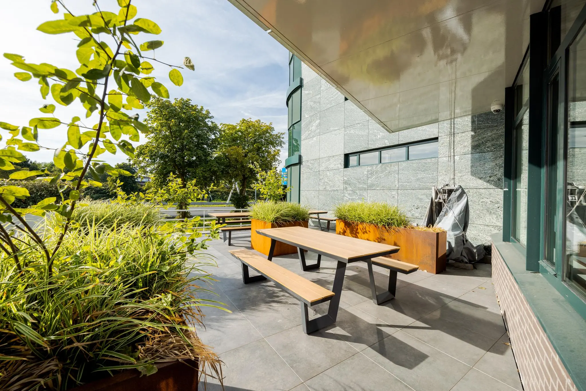 Zonnig terras met houten picknicktafels en groene planten naast een modern kantoorgebouw aan de Euclideslaan.