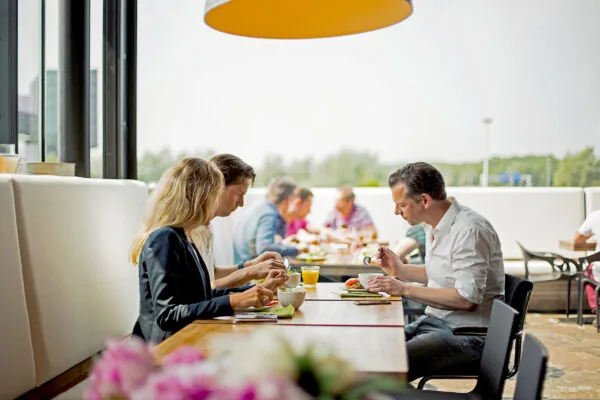 Mensen eten en praten samen aan een lange tafel in een open en modern bedrijfsrestaurant aan de Orteliuslaan.