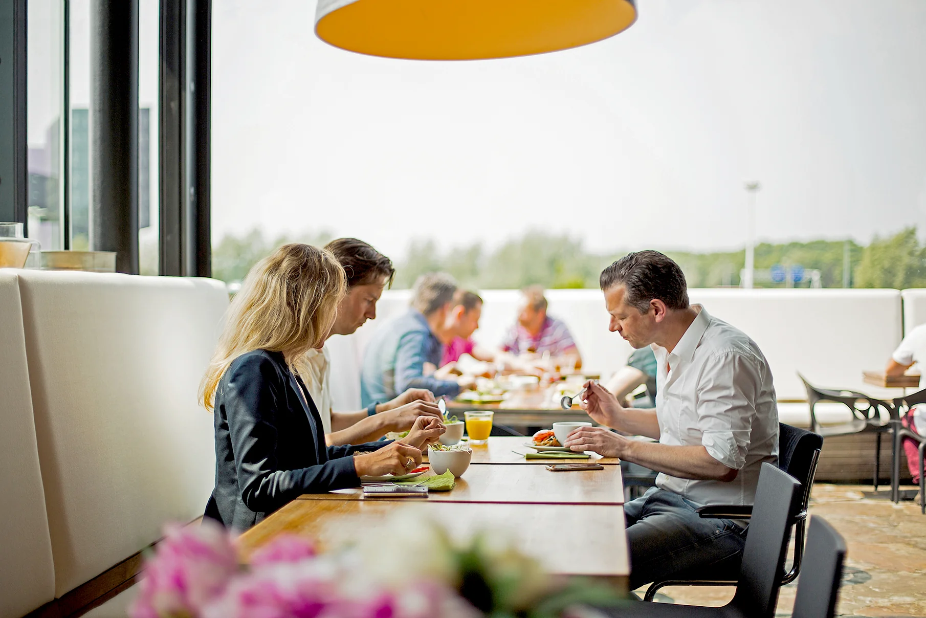 Mensen eten en praten samen aan een lange tafel in een open en modern bedrijfsrestaurant aan de Orteliuslaan.