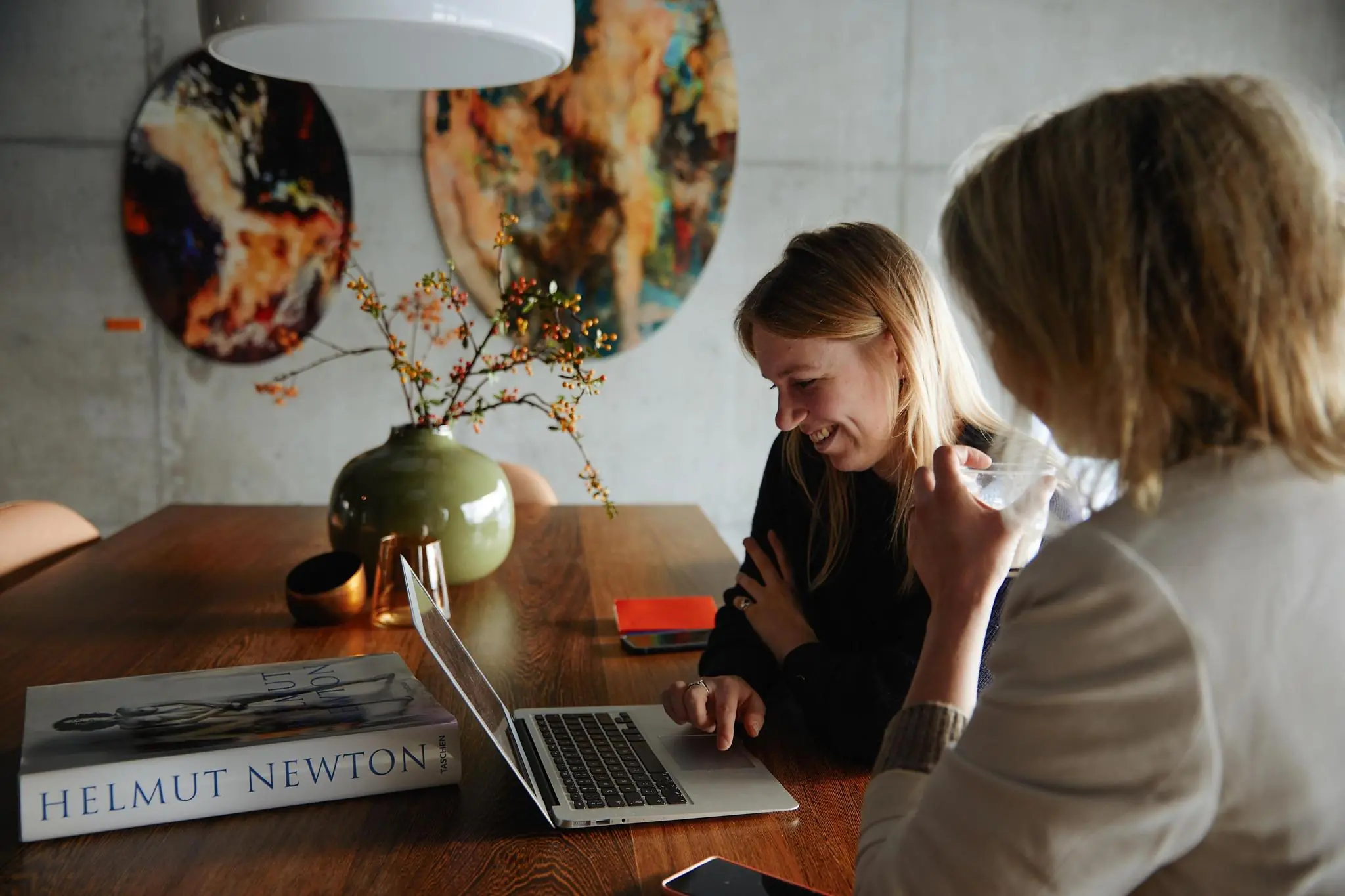 Twee vrouwen zitten samen aan een houten tafel in een modern interieur op Orteliuslaan en kijken lachend naar een laptop.