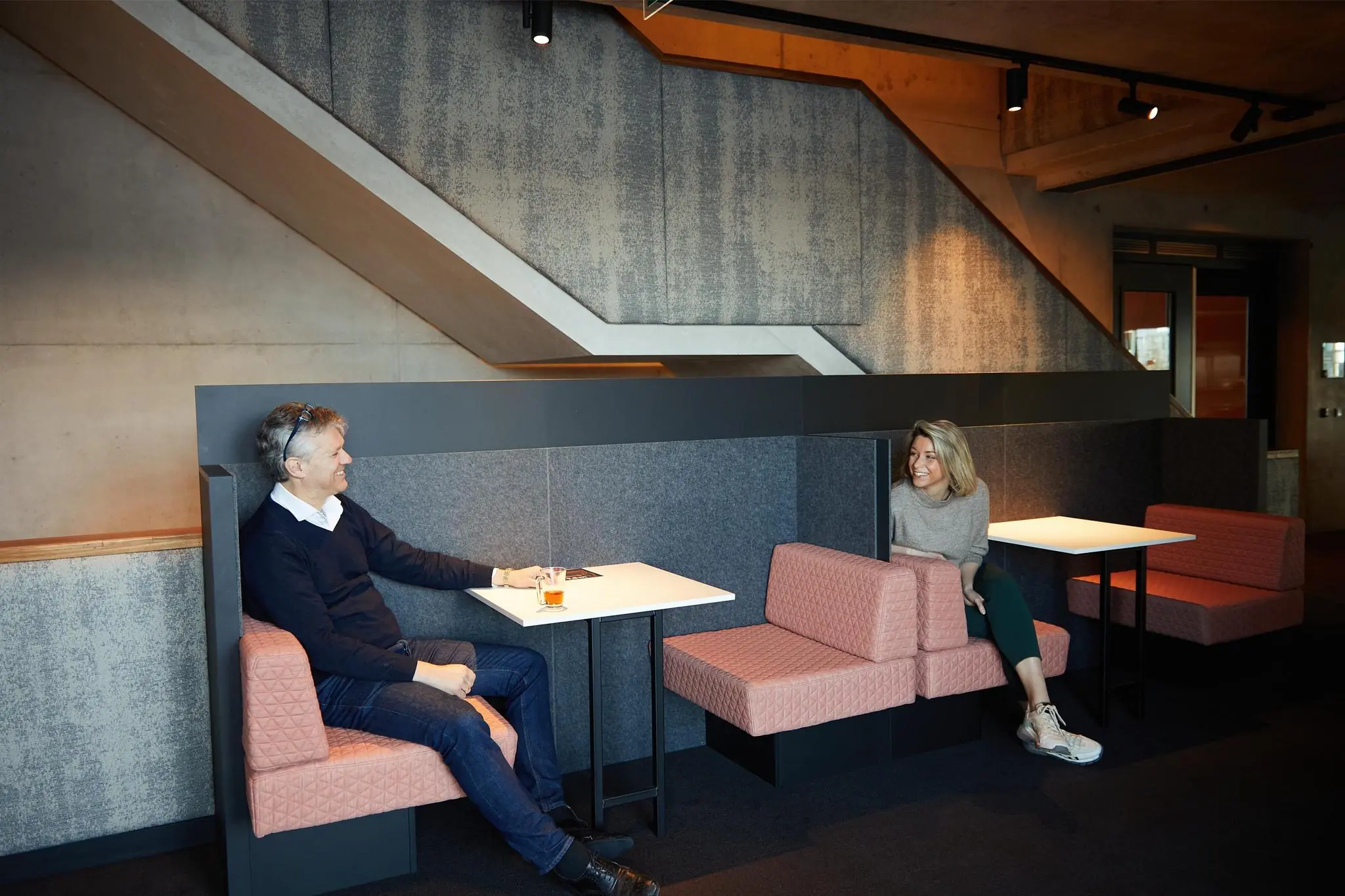 Twee mensen zitten tegenover elkaar in een moderne zitruimte op Orteliuslaan, met betonnen muren en roze gestoffeerde banken.
