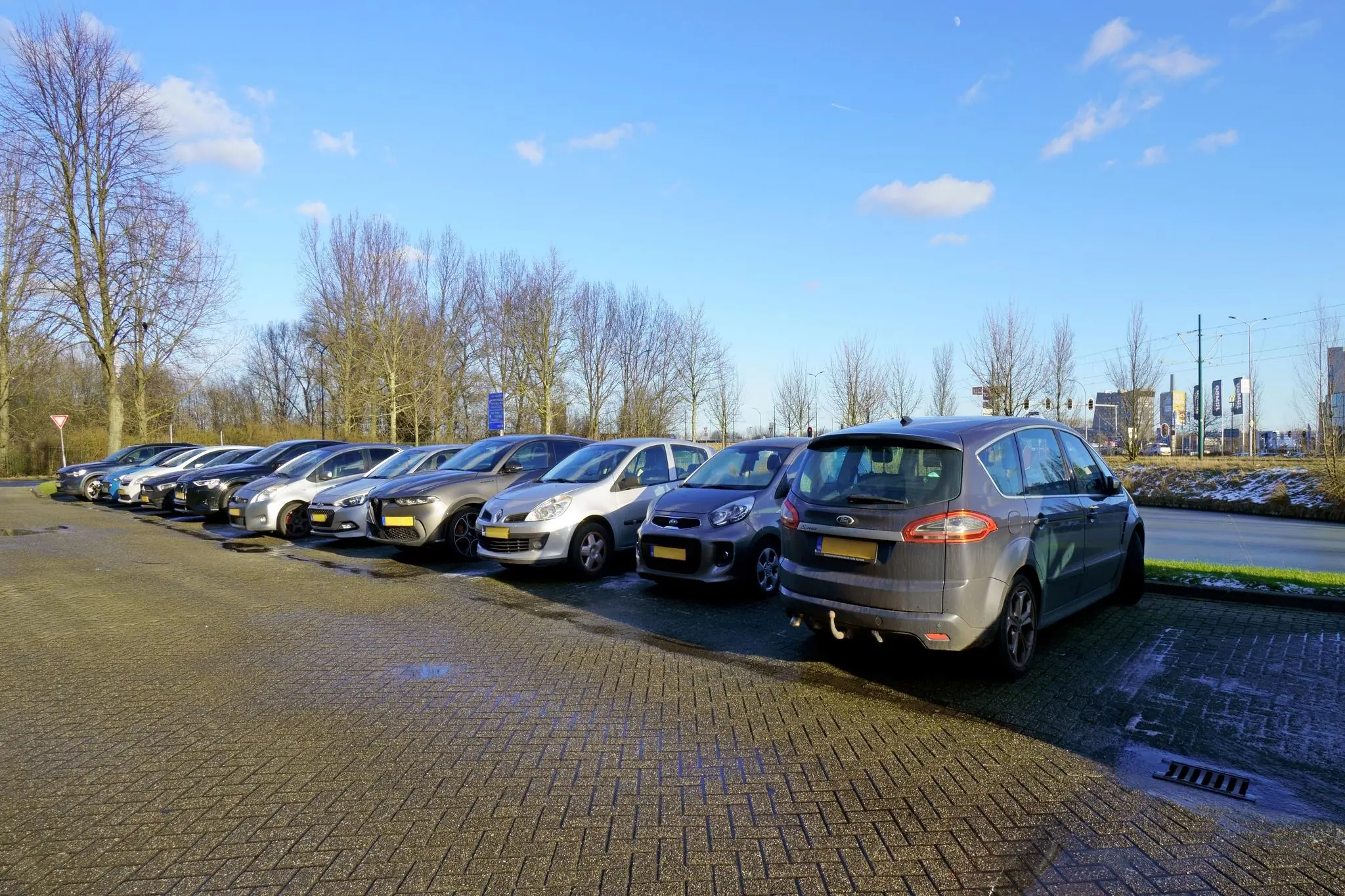 Rij geparkeerde auto’s op een deels nat parkeerterrein aan de Zuiderweg, met kale bomen en een heldere blauwe lucht op de achtergrond.