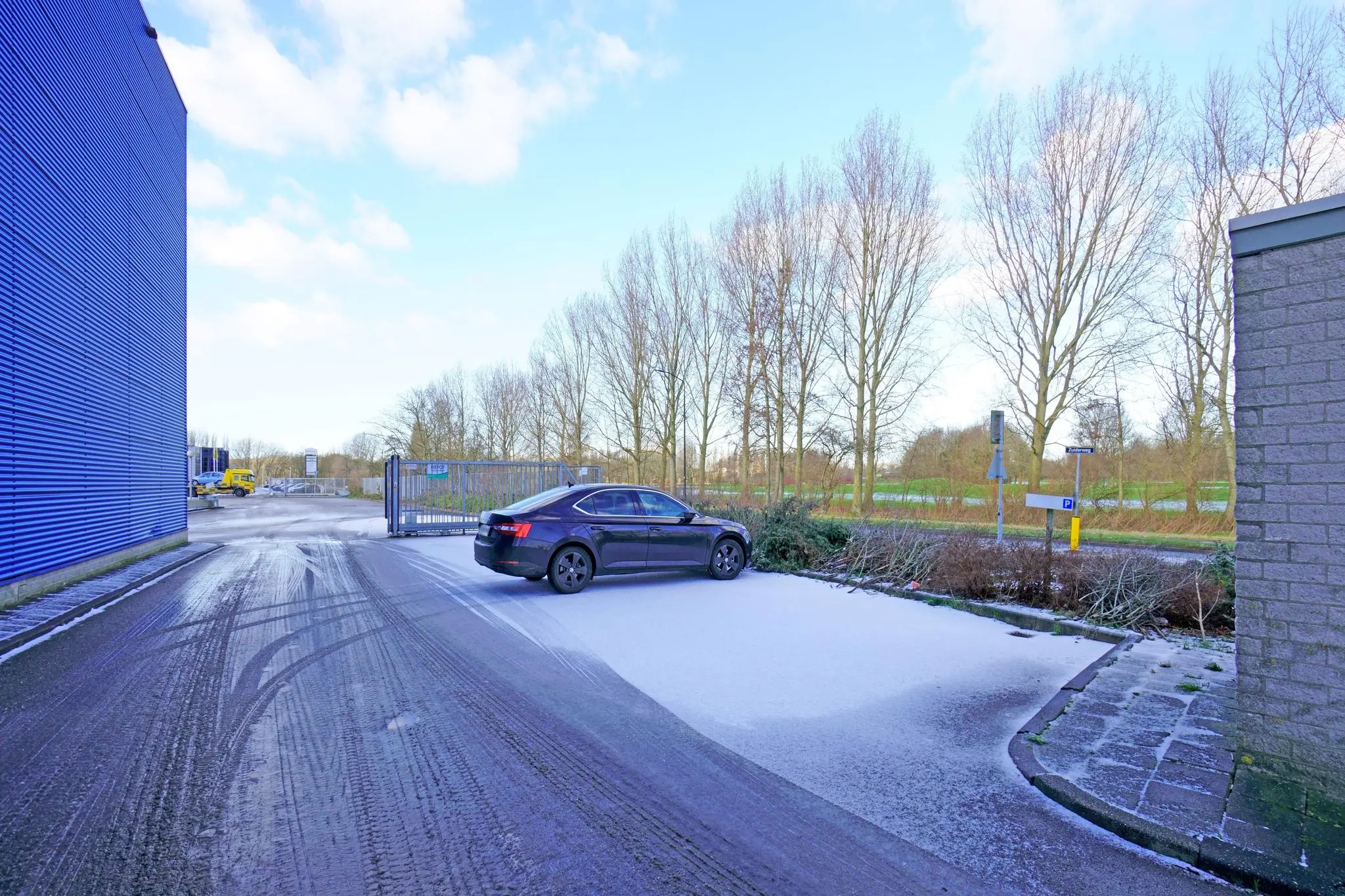 Zwarte auto geparkeerd op besneeuwd terrein langs de Zuiderweg bij een bedrijventerrein met blauwe gevel en toegangspoort.