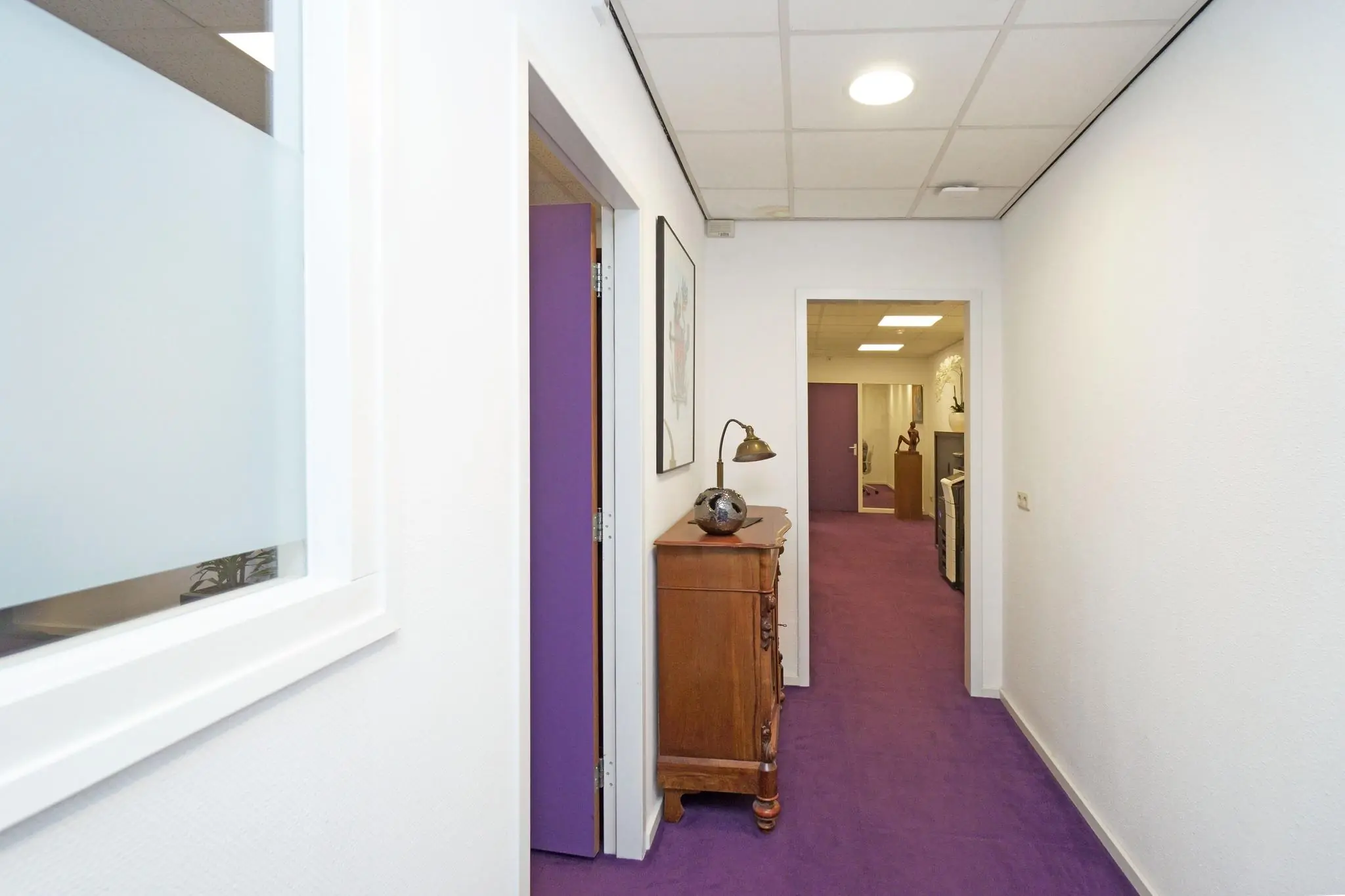 Witte gang met paarse vloerbedekking en een houten kast met lamp, in een kantoor aan de Zuiderweg.