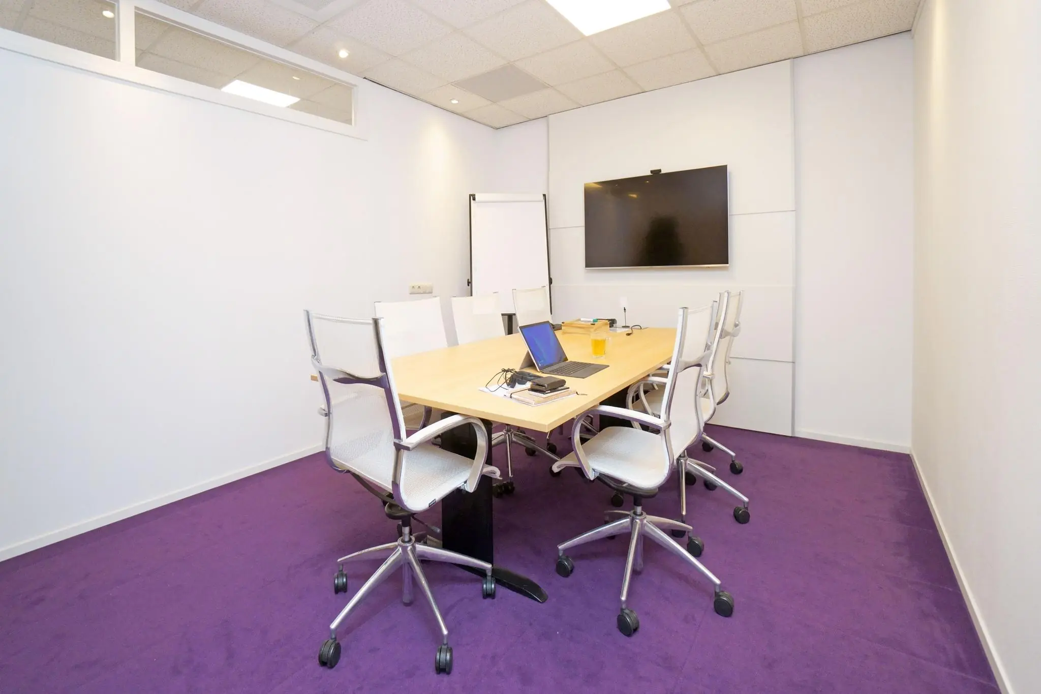 Vergaderruimte aan de Zuiderweg met paarse vloerbedekking, een vergadertafel, acht stoelen, een laptop, een flatscreen en een flip-over.