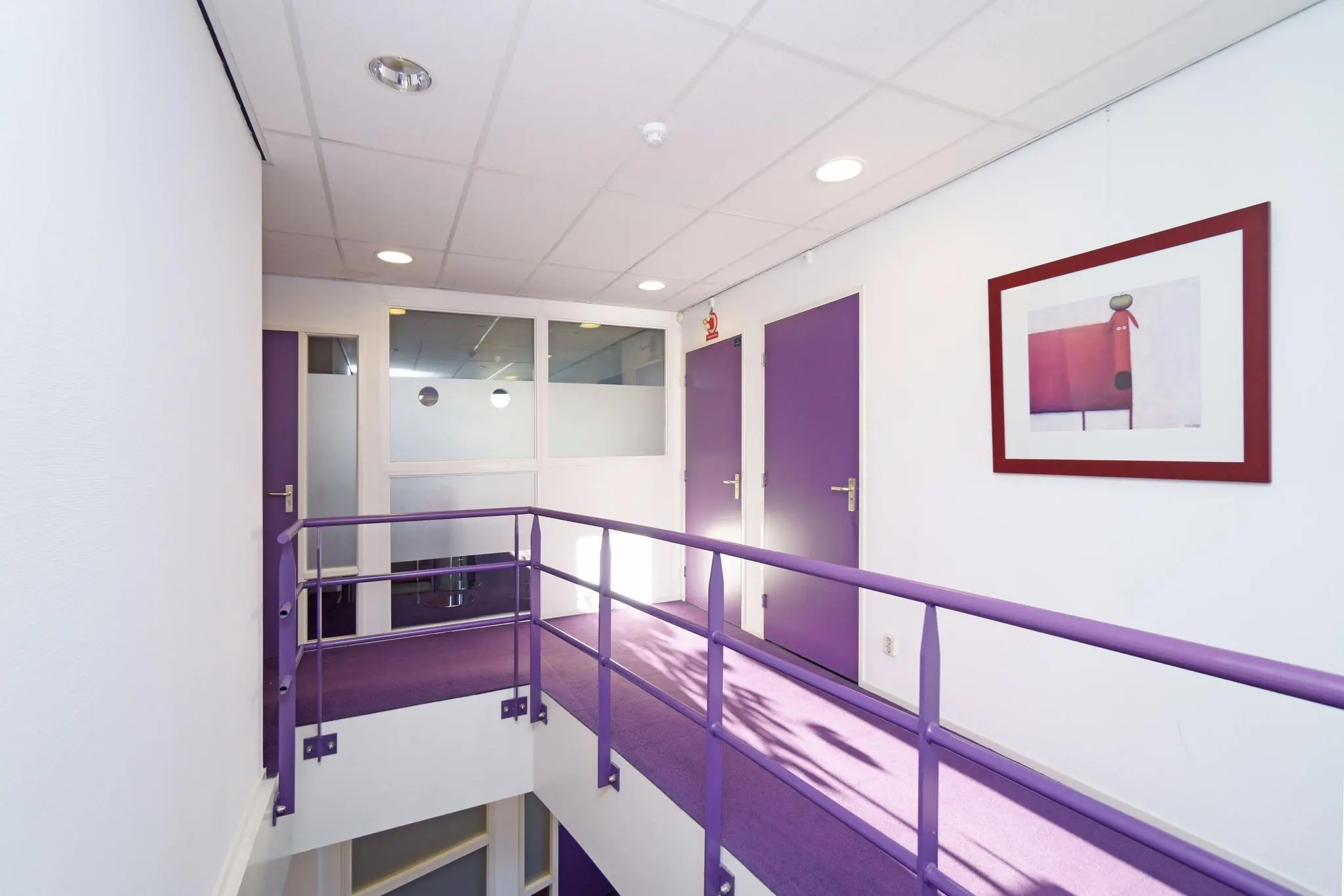 Gang op een bovenverdieping met paarse deuren, paarse vloerbedekking en een kunstwerk aan de witte muur.
