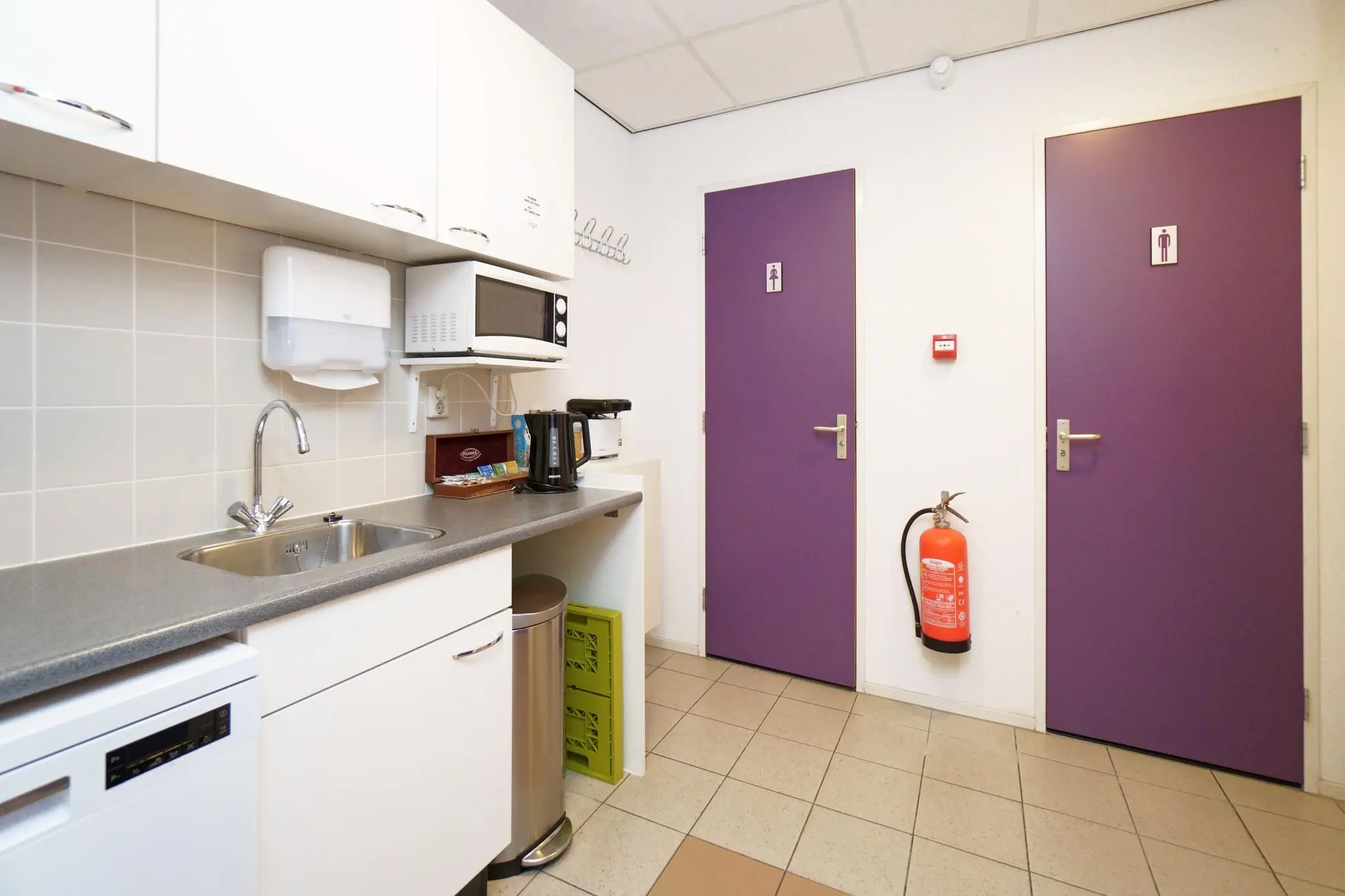 Kleine kitchenette met paarse toiletdeuren, koffiezetapparaat, magnetron en een brandblusser aan de muur.